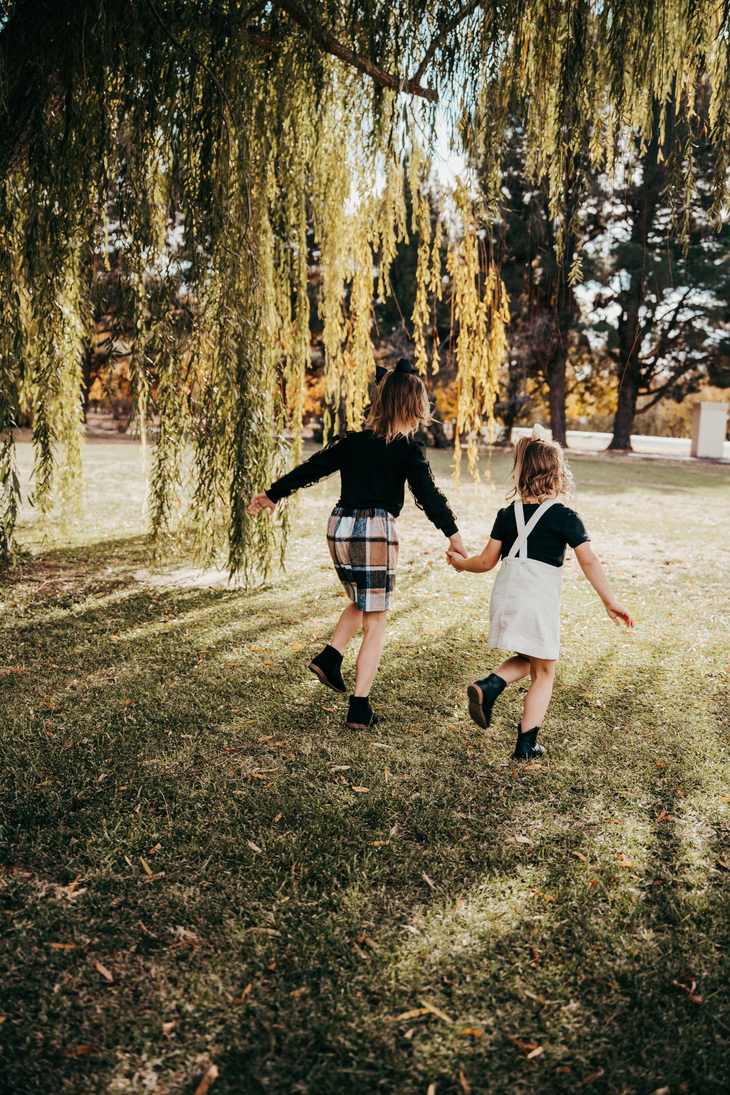 Two girls holding hands running through a park under a weeping willow tree in autumn.