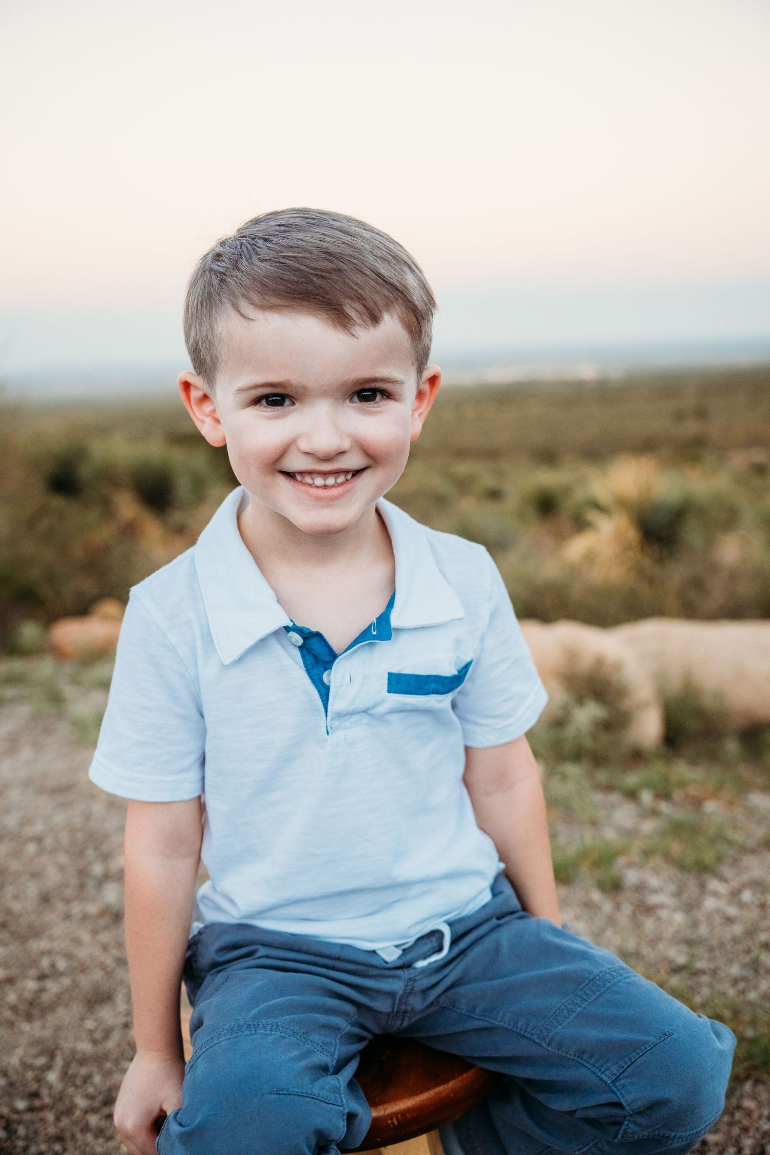 Smiling young boy in a light blue polo shirt sitting outdoors with a natural landscape behind him.