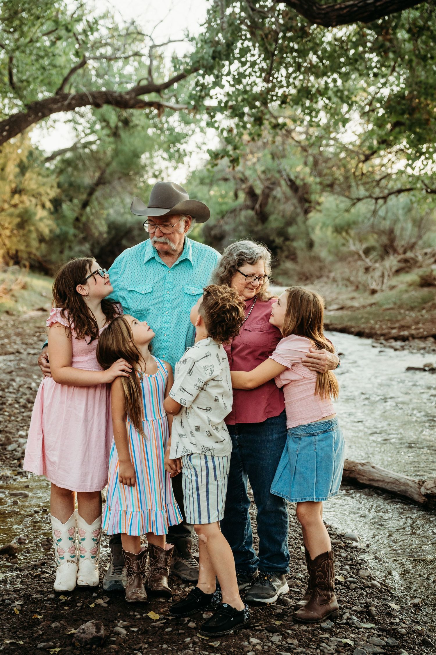 Grandparents share a warm group hug with four grandchildren beside a scenic tree-lined creek.