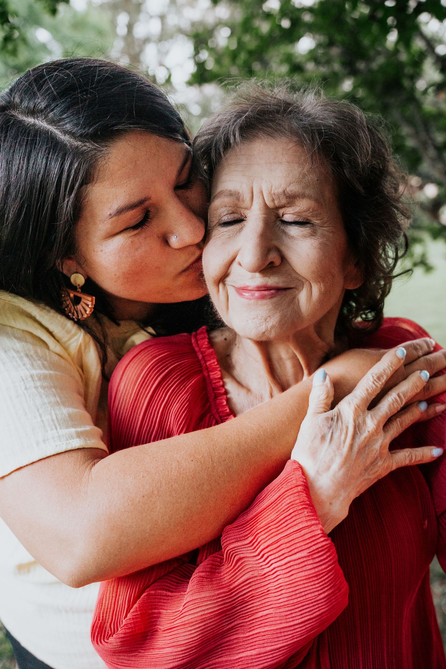 A woman tenderly kisses and hugs an elderly woman in a red blouse in an outdoor setting.