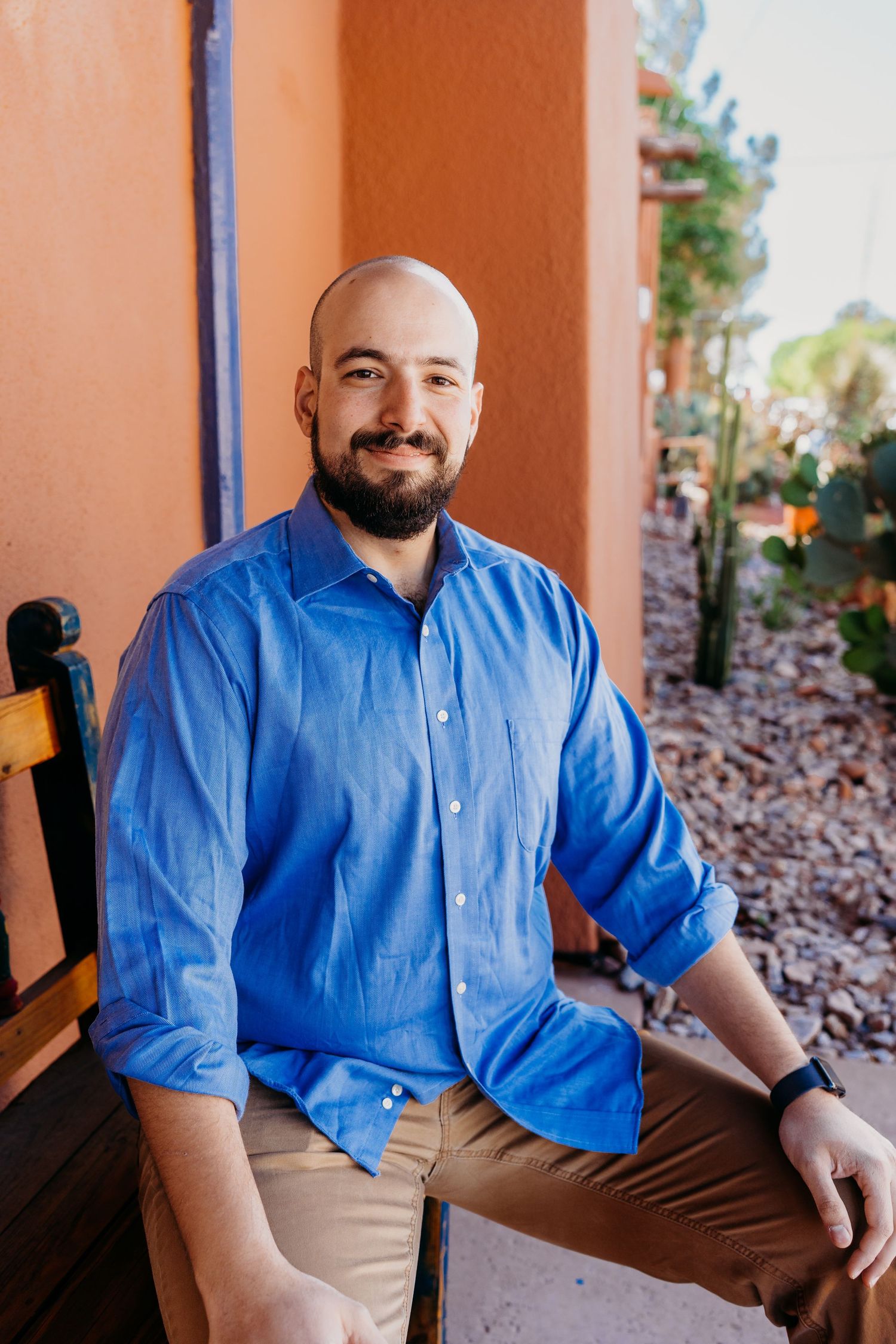 Bearded bald man in blue shirt sitting on bench beside desert rock garden.