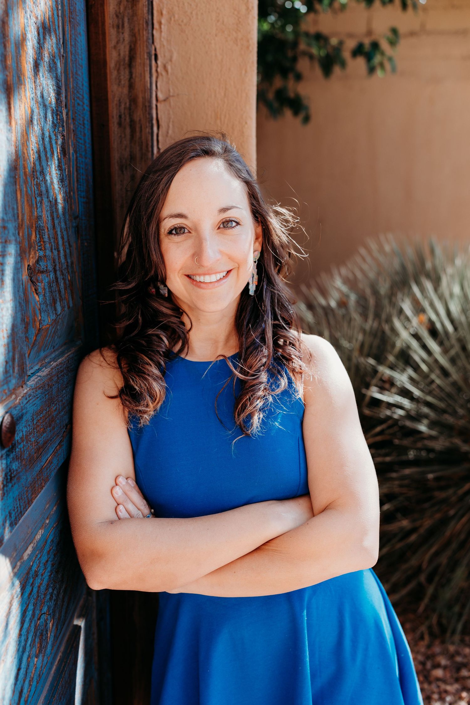 Cheerful brunette woman in blue dress with arms crossed leaning on wooden door.
