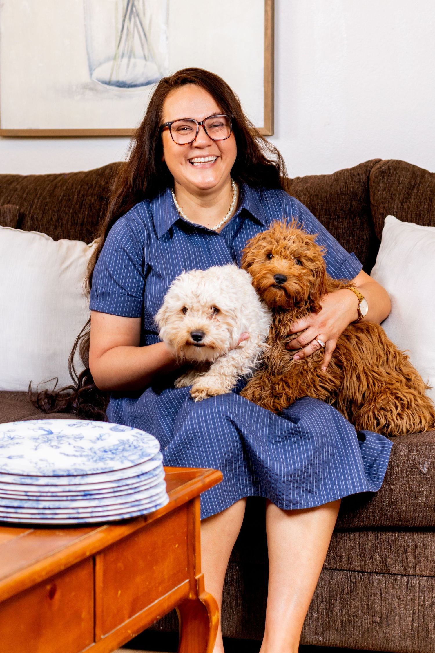 Smiling woman in blue dress holding two fluffy dogs, white and brown, on a brown sofa.