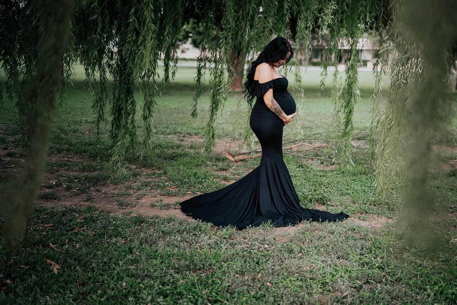 Pregnant woman in a black mermaid maternity gown standing gracefully under weeping willow branches.