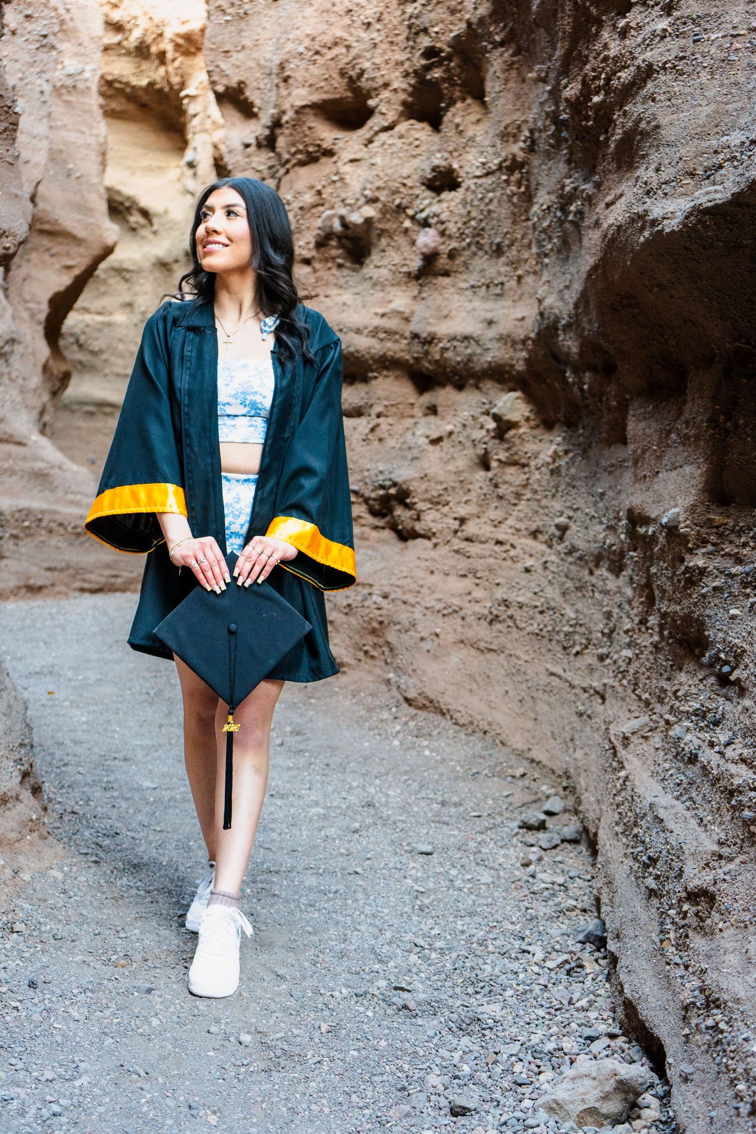 Female graduate in black and gold gown carrying cap while strolling through a sandy canyon.
