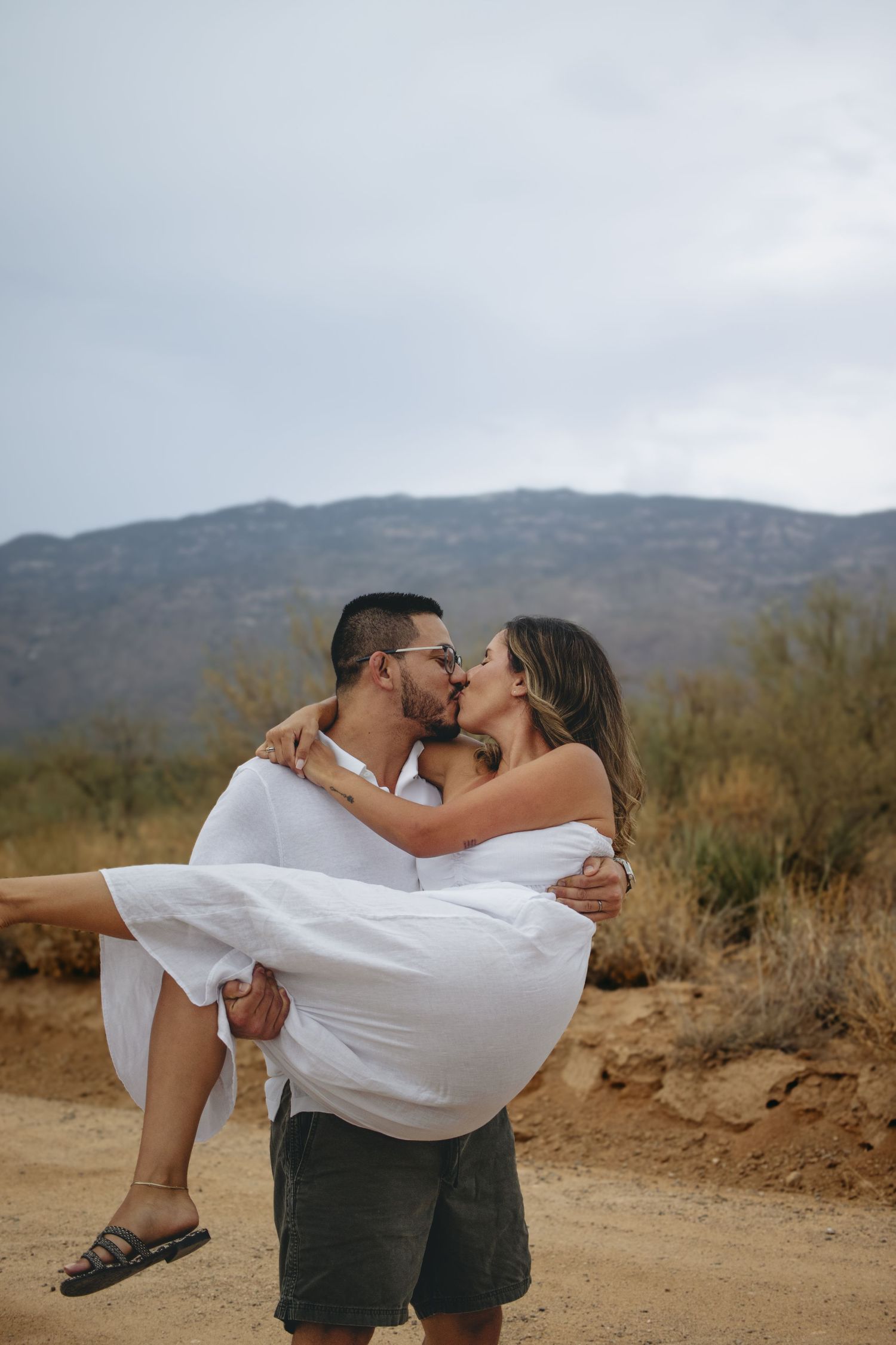 Man carrying woman in white dress while sharing a kiss on a desert dirt road.