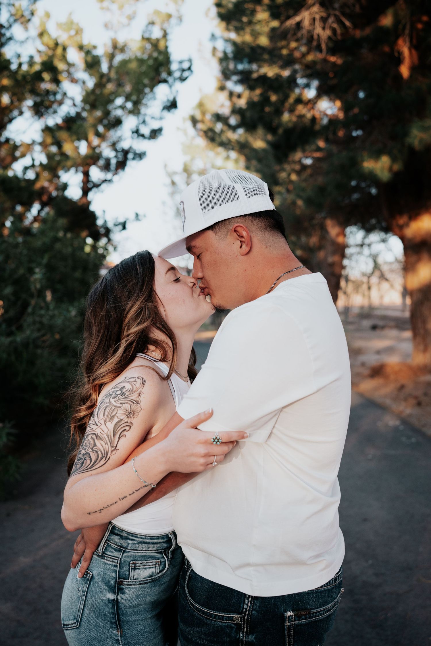 Tattooed couple sharing a romantic kiss outdoors, man wearing white cap and t-shirt, woman in jeans.