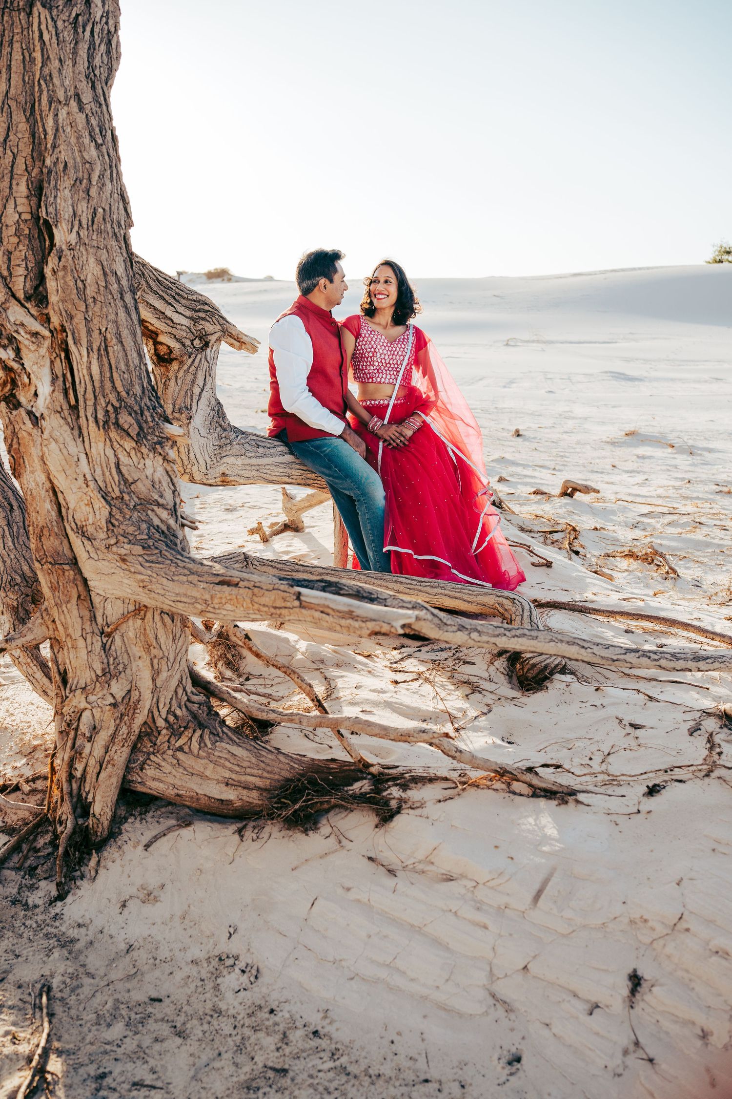 Indian couple in vibrant red traditional outfits laughing together on driftwood in white sands.
