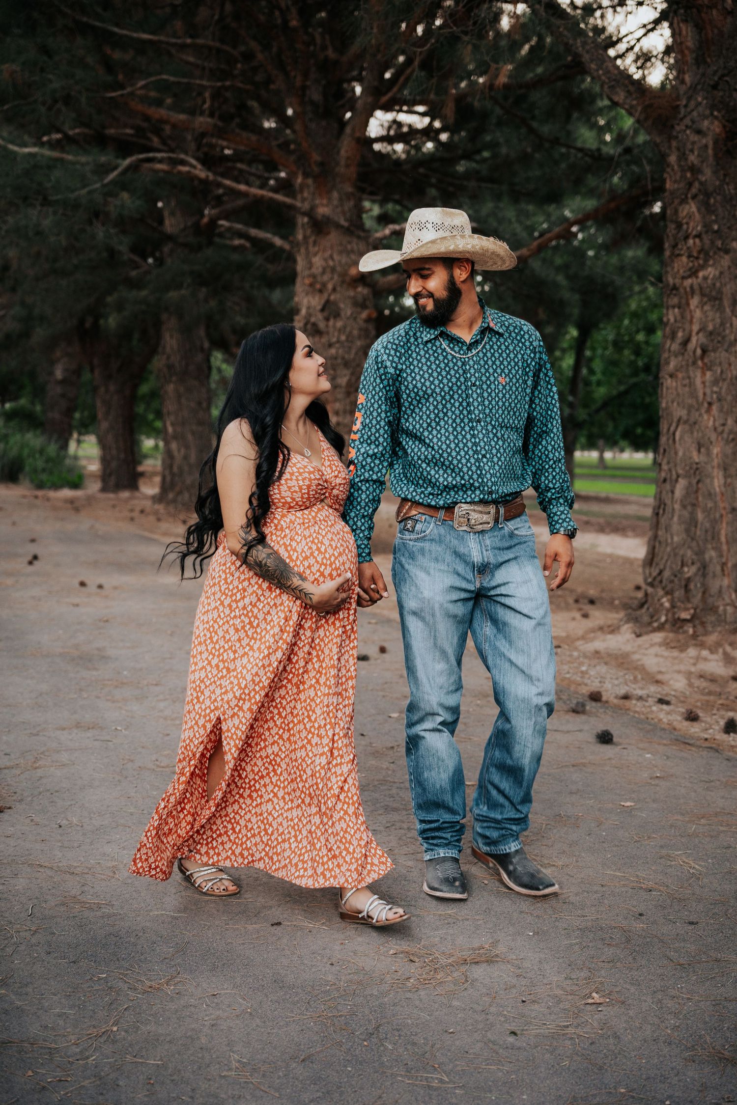 Pregnant woman in orange floral maxi dress walks with cowboy partner in teal shirt and straw hat.