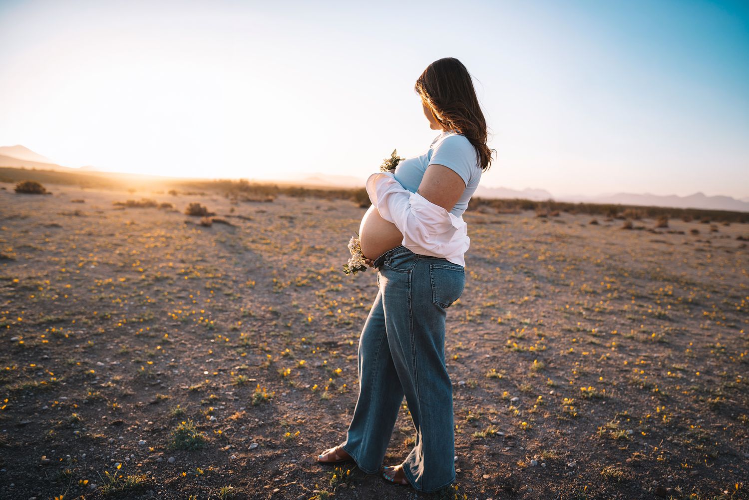 Pregnant woman holds baby bump and wildflowers in open desert at golden sunset.