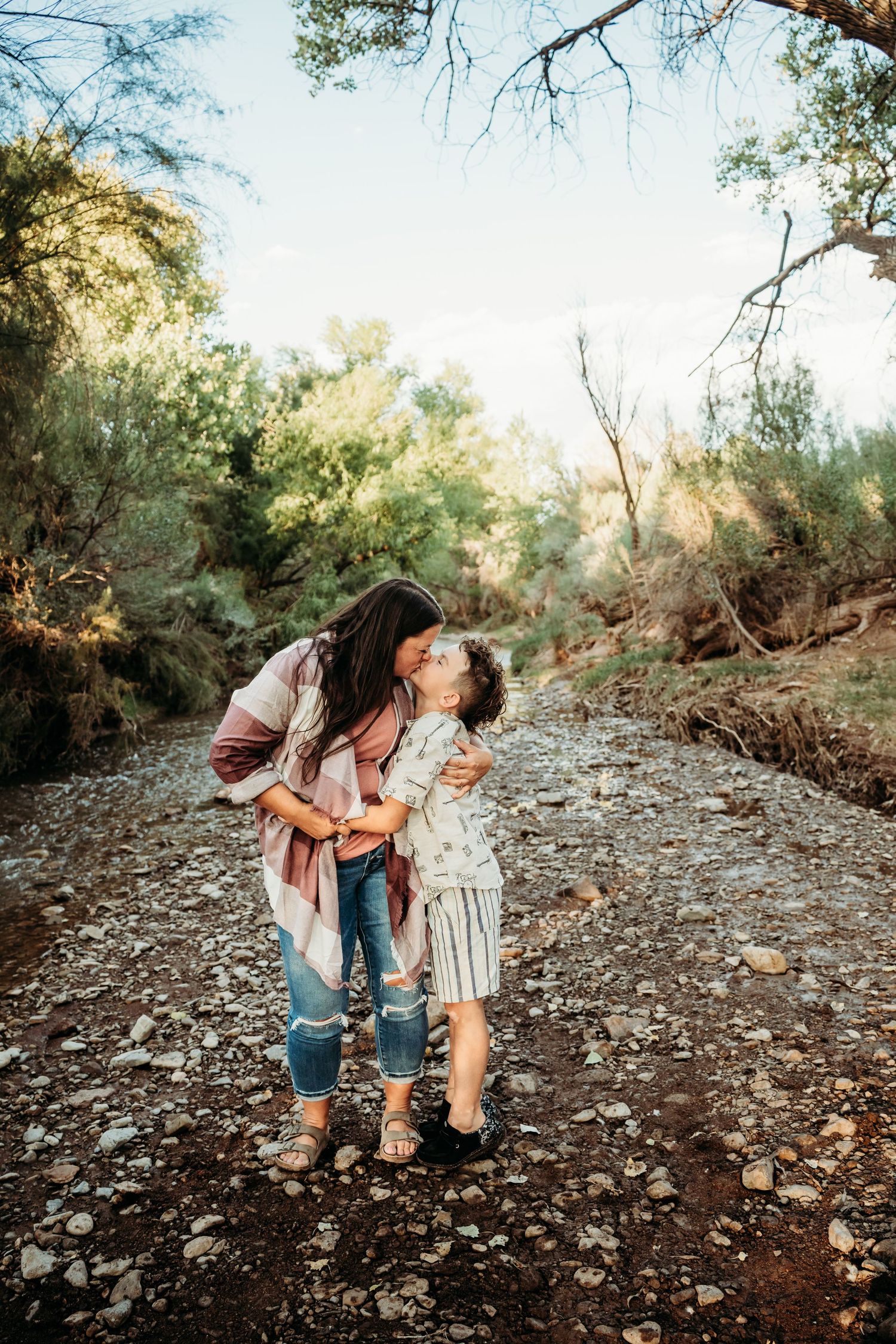 A loving mother leans down to kiss her young son while standing on a rocky creekbed among trees.