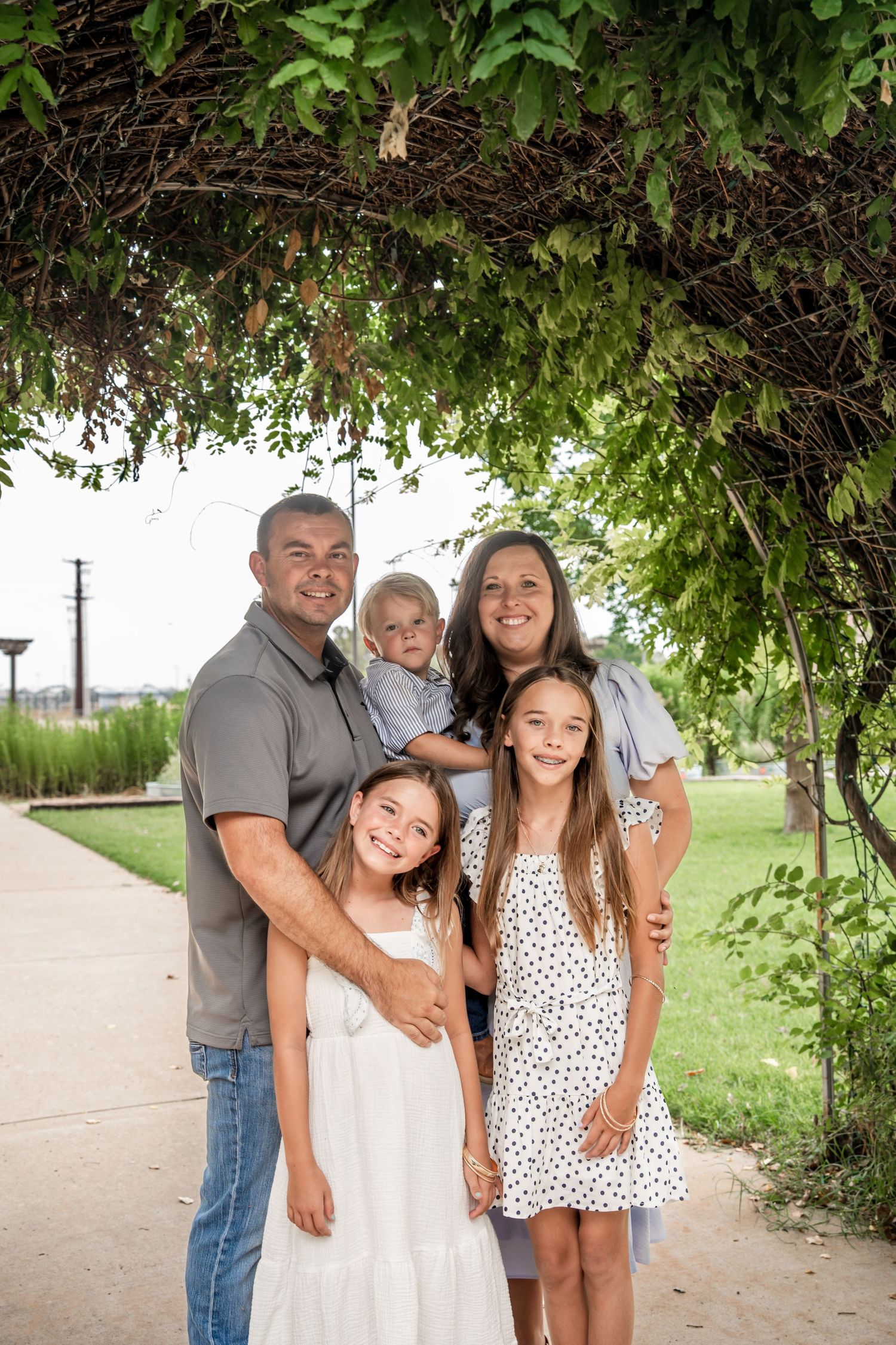 Family of five smiling under leafy green archway in outdoor park setting.