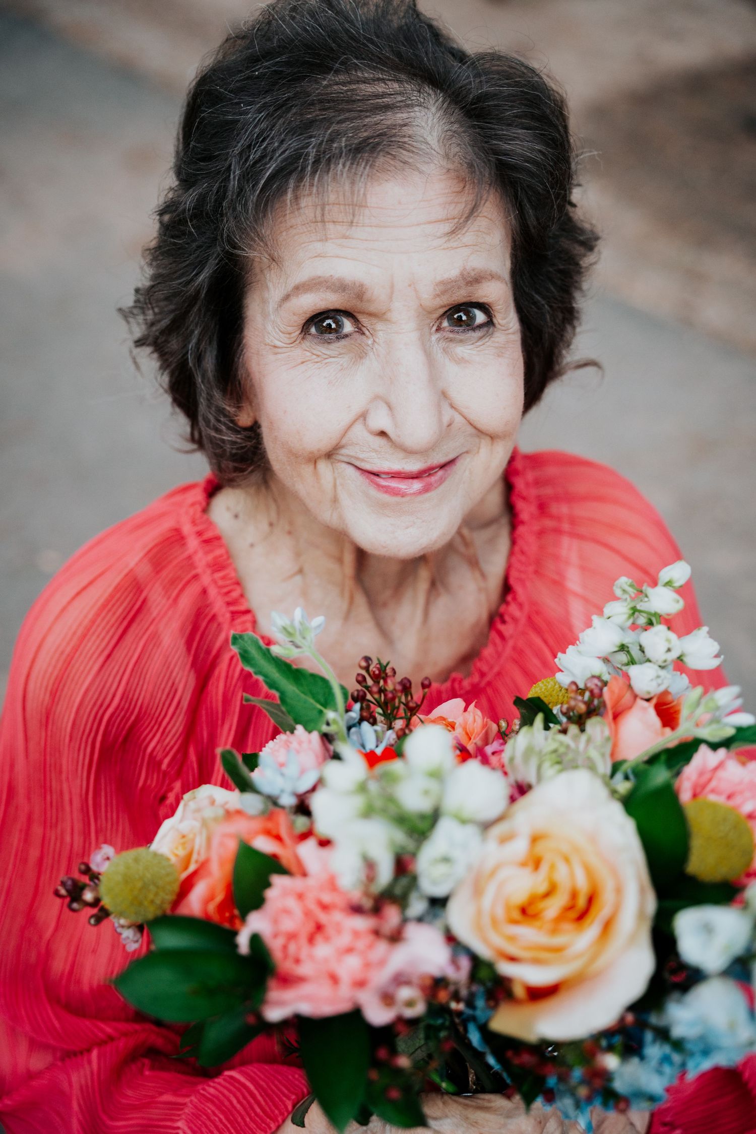Elderly woman in red blouse smiles warmly while holding a colorful bouquet of roses and flowers.