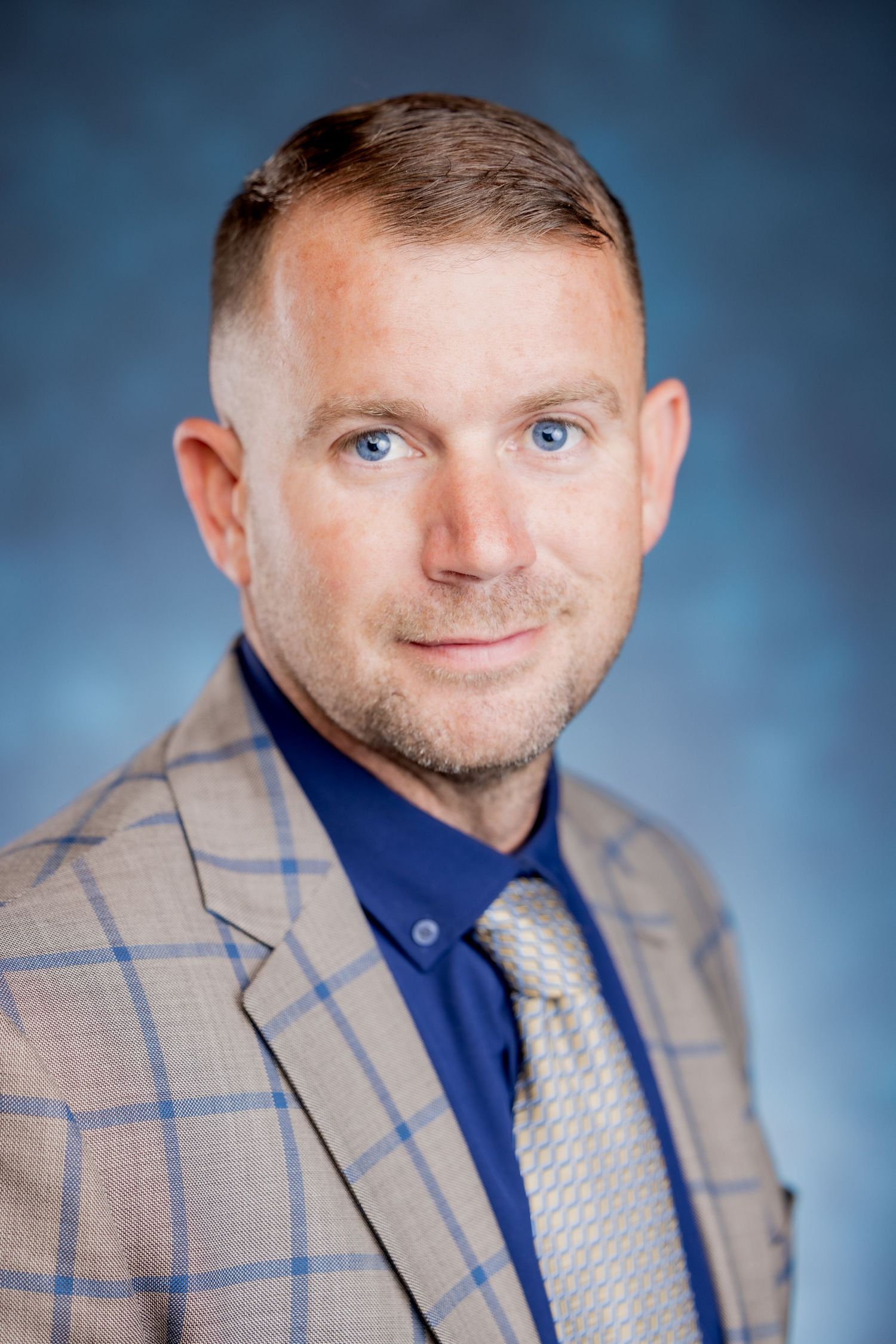Tight professional headshot of a blue-eyed man in a tan plaid blazer and navy shirt on blue background.