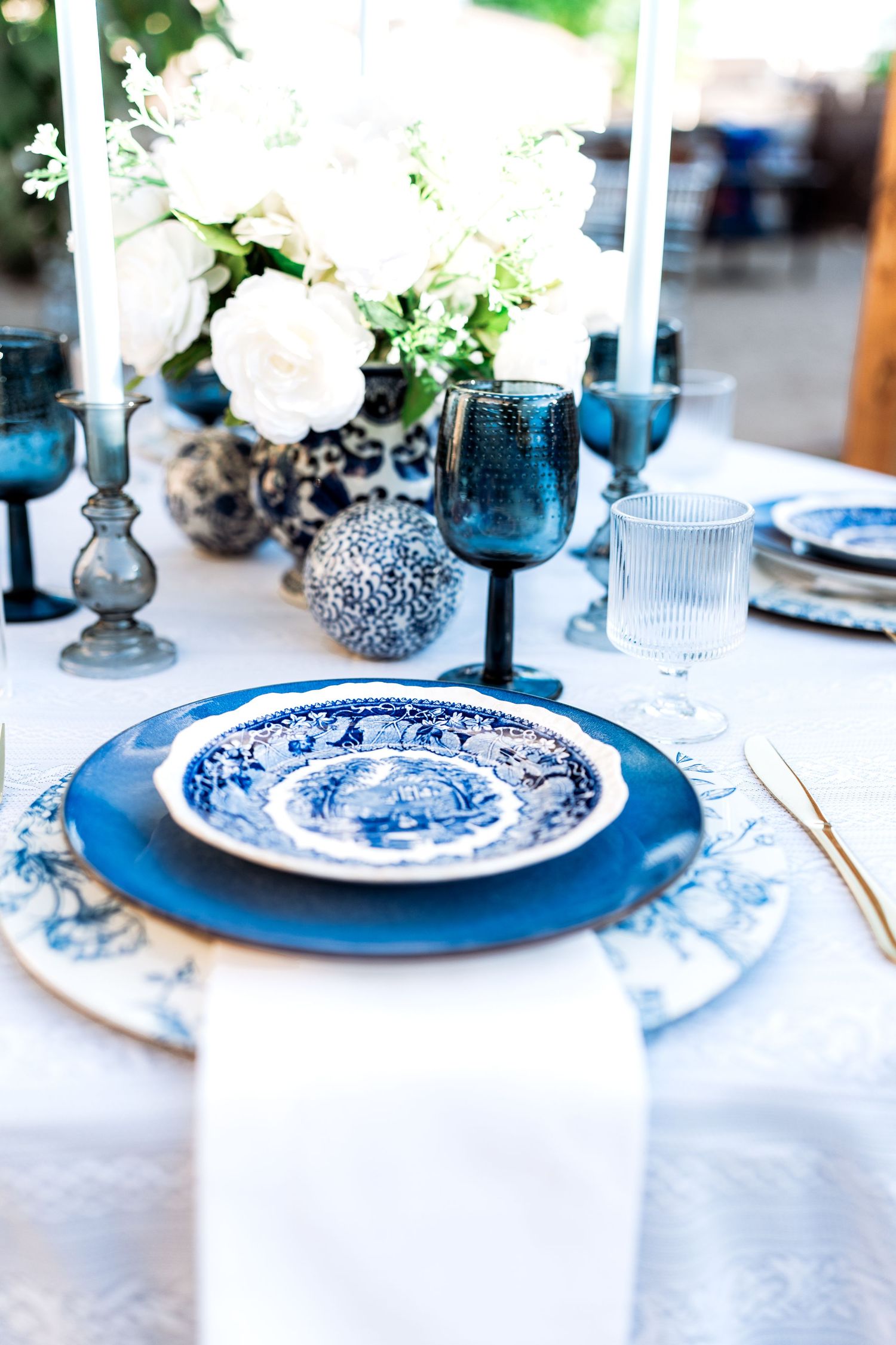 Blue and white wedding tablescape featuring layered china plates, dark blue goblets, and white rose centerpiece.
