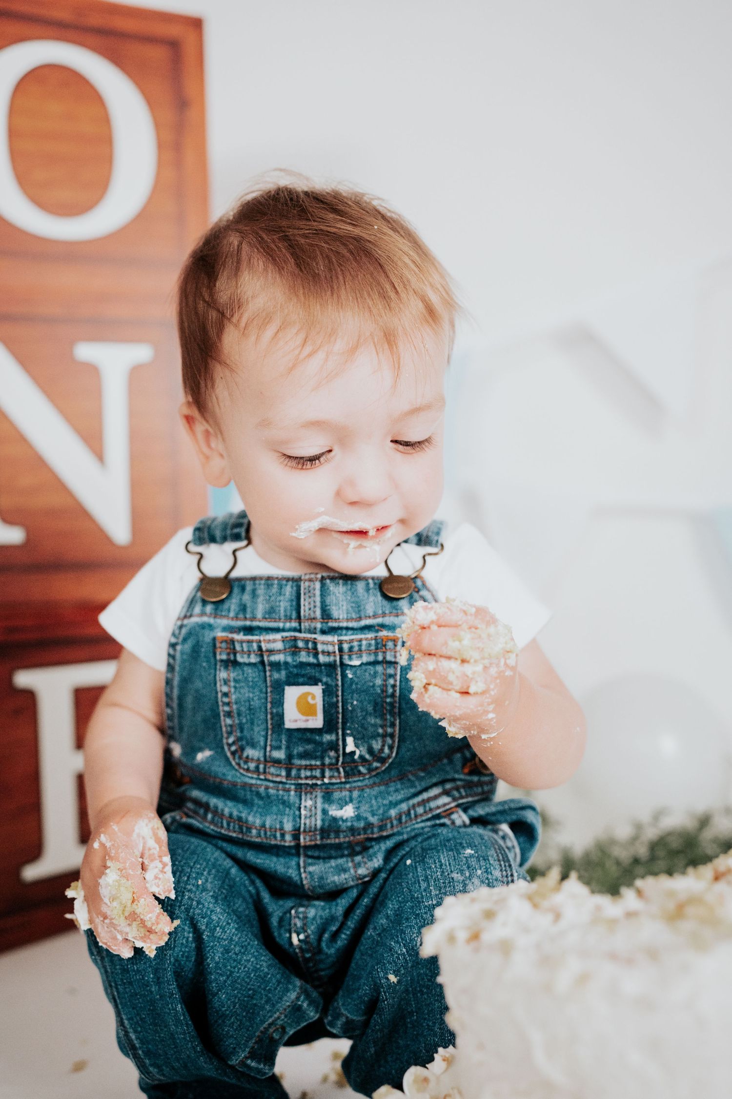 Sweet baby boy in denim overalls examining cake frosting on his hands near ONE birthday sign.