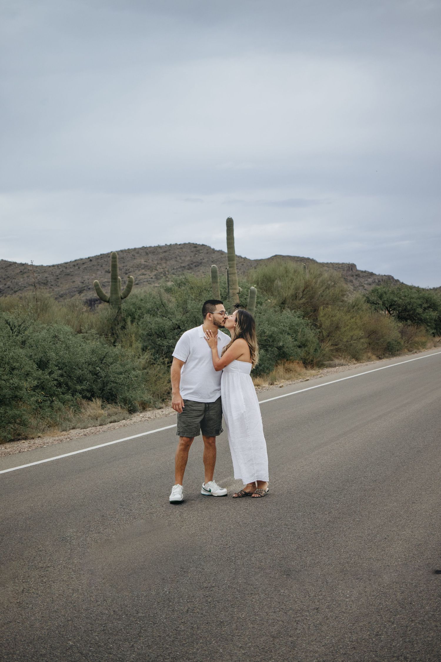 Couple kissing on empty desert road surrounded by saguaro cacti and desert landscape.