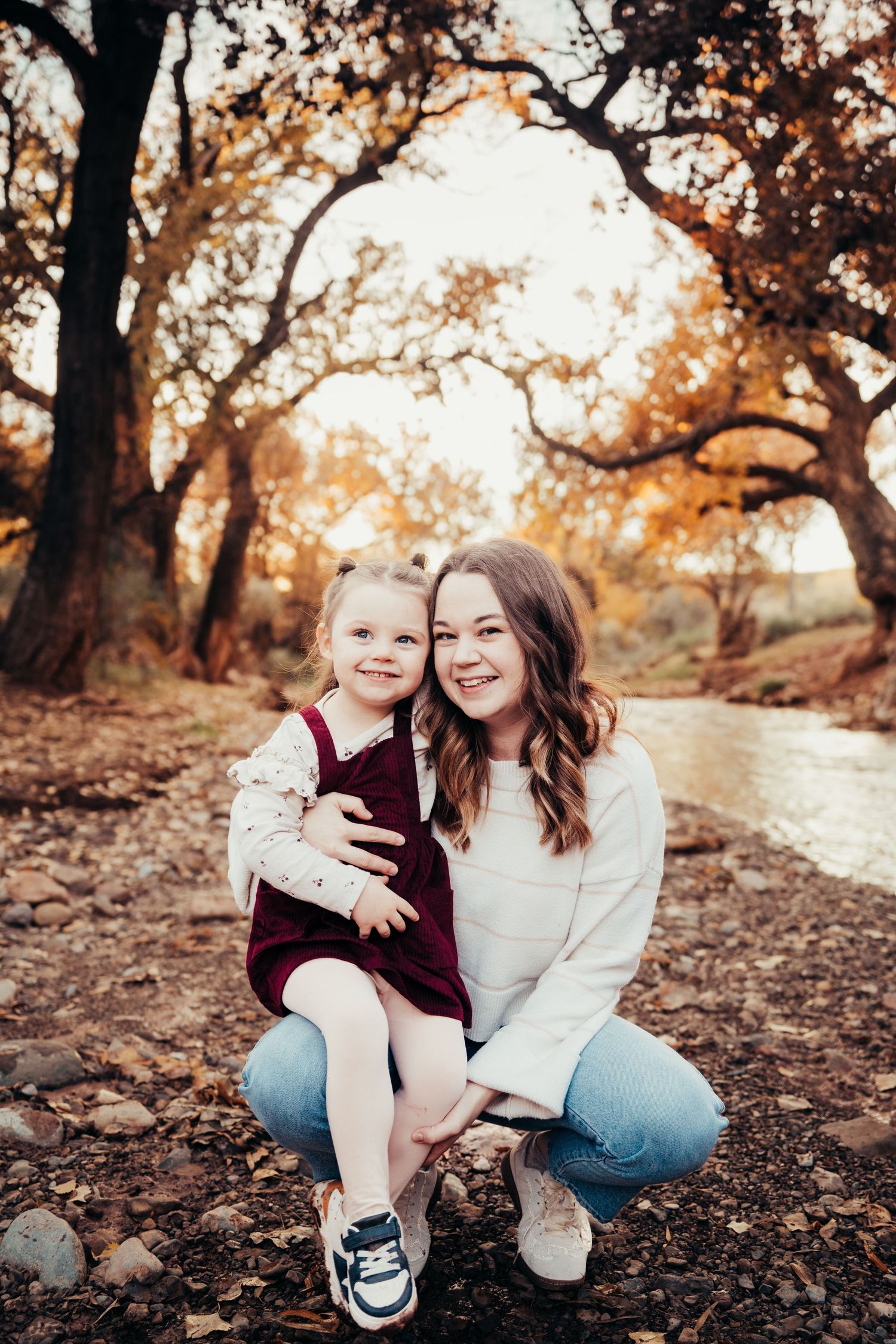 Smiling mother crouching with happy toddler daughter in burgundy dress by a creek in autumn forest.
