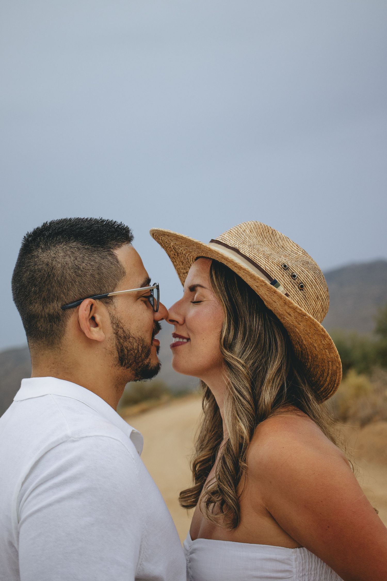 Couple sharing a romantic nose-to-nose moment outdoors, woman wearing straw hat and white dress.