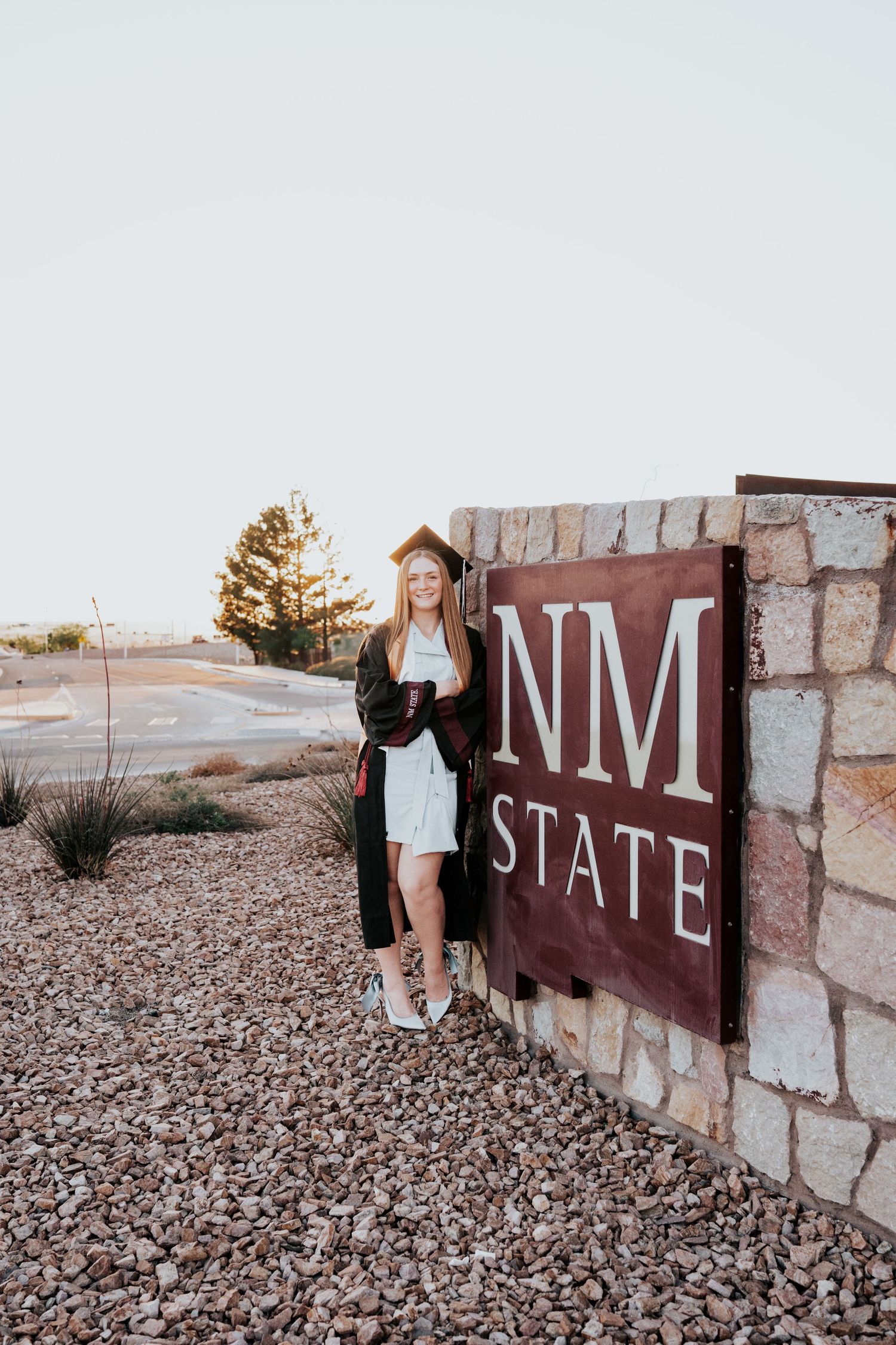 Blonde female graduate in cap and gown posing beside NM State stone entrance sign.