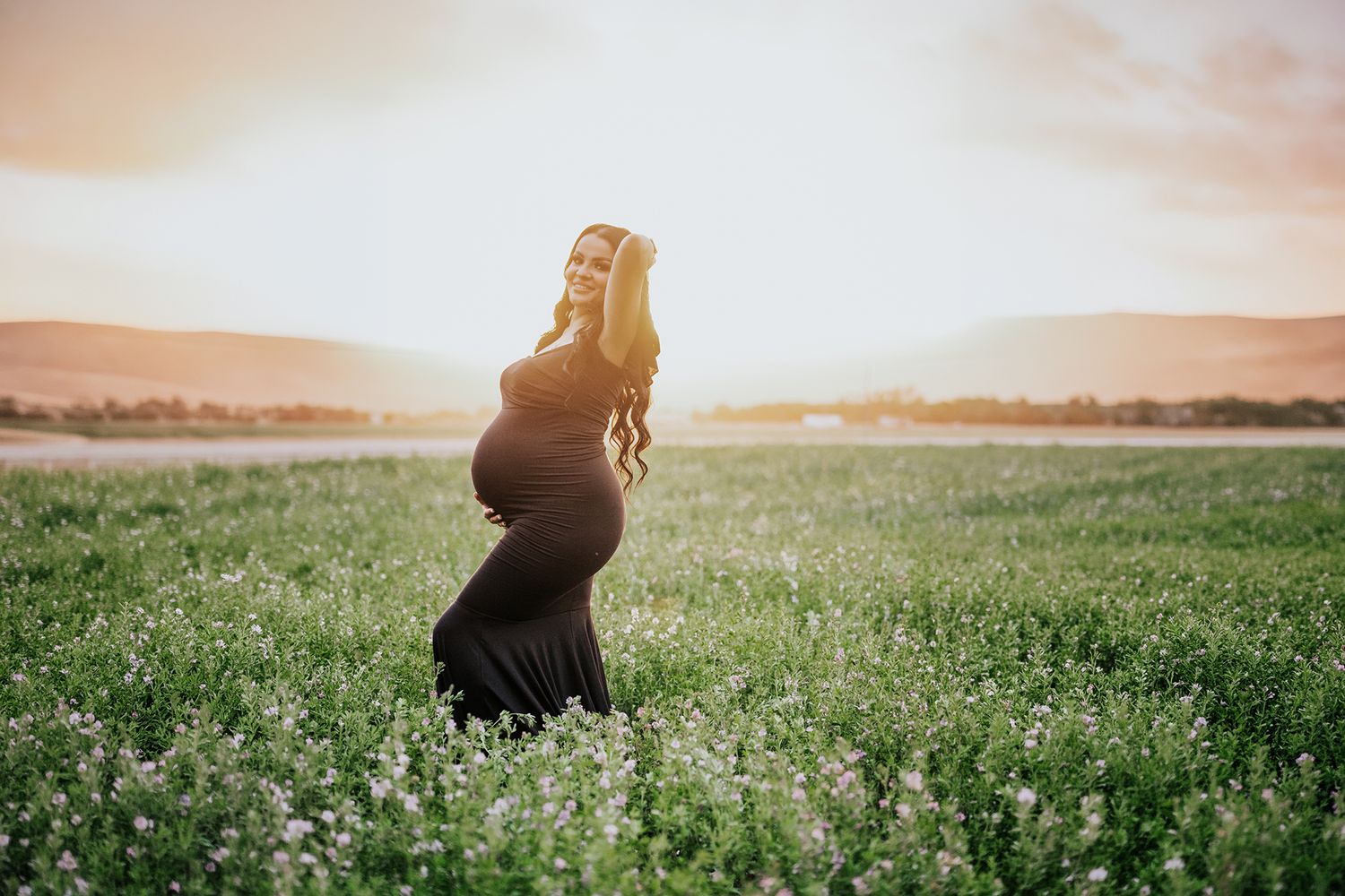 Pregnant woman in black gown holding belly in flower field during golden sunset.