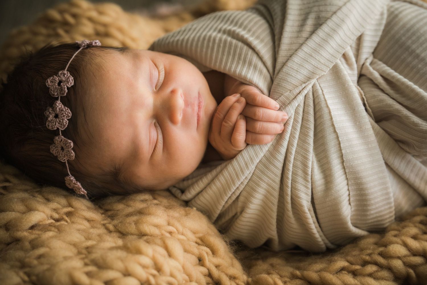 Close-up of sleeping newborn baby girl in striped swaddle with floral headband on chunky knit blanket.