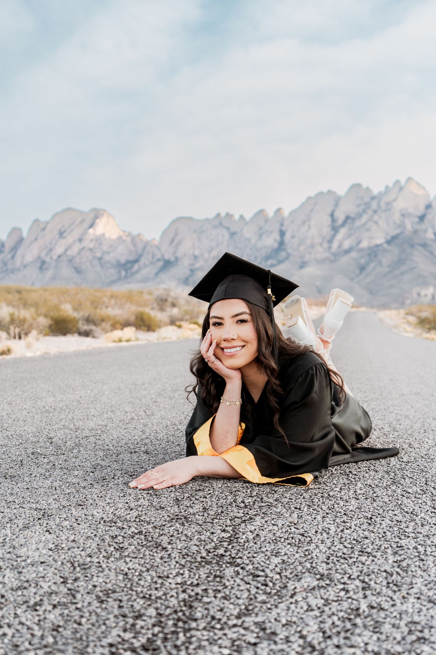 Smiling female graduate in black cap and gold gown lies on a desert road with scenic mountain backdrop.