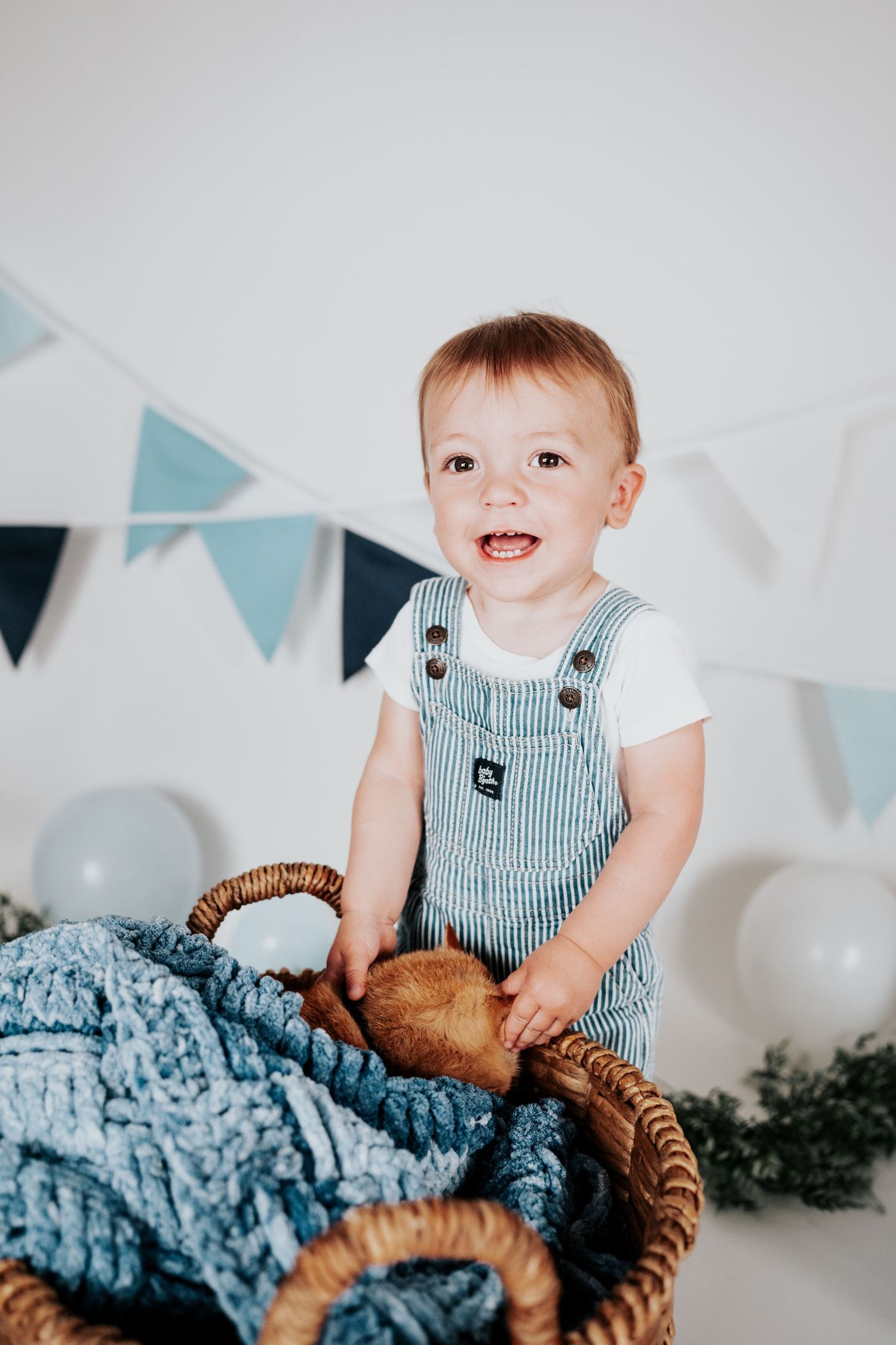 Happy baby boy in blue striped overalls laughing while standing in a large wicker basket with festive decor.