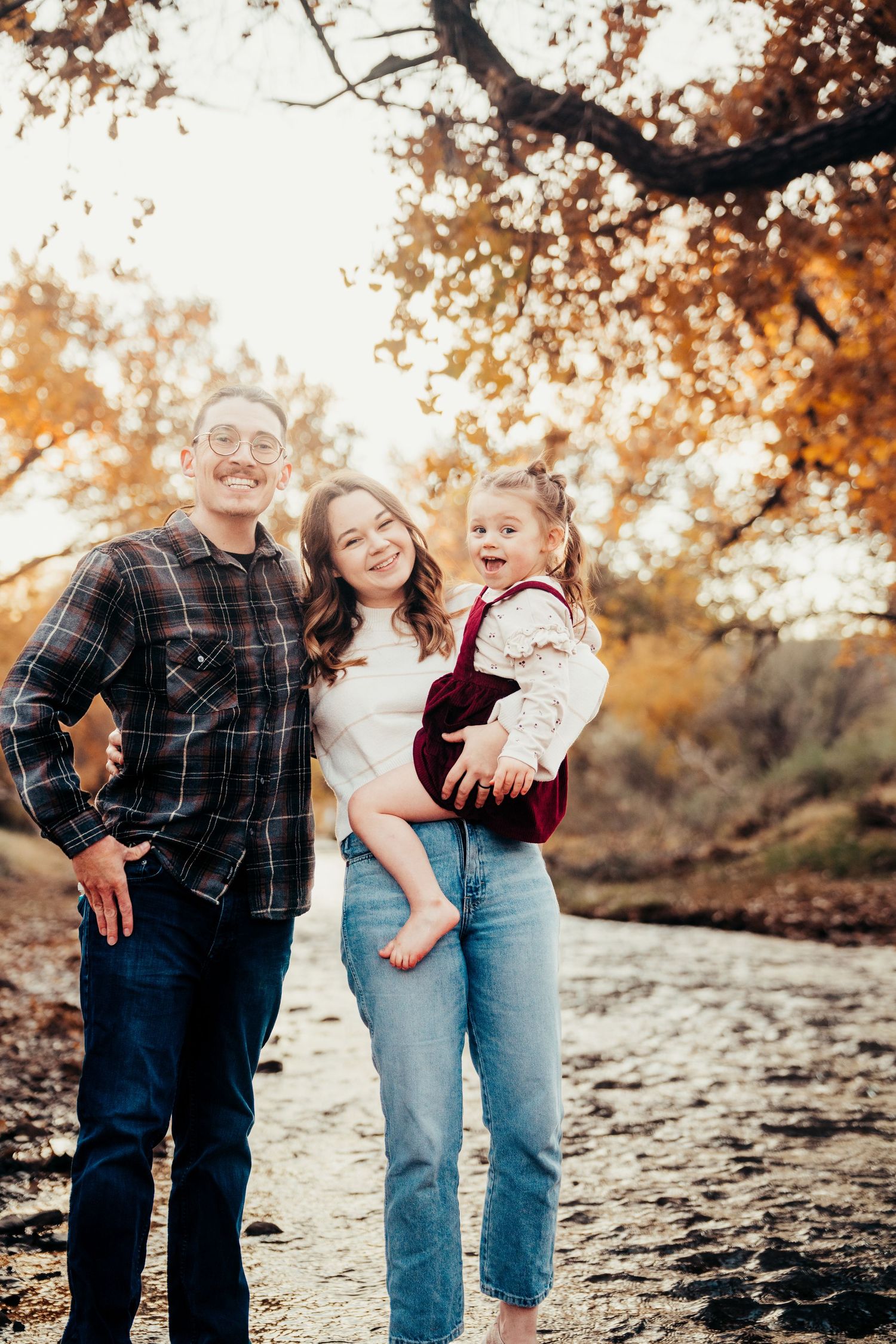 Happy family of three poses together near creek surrounded by golden fall foliage.