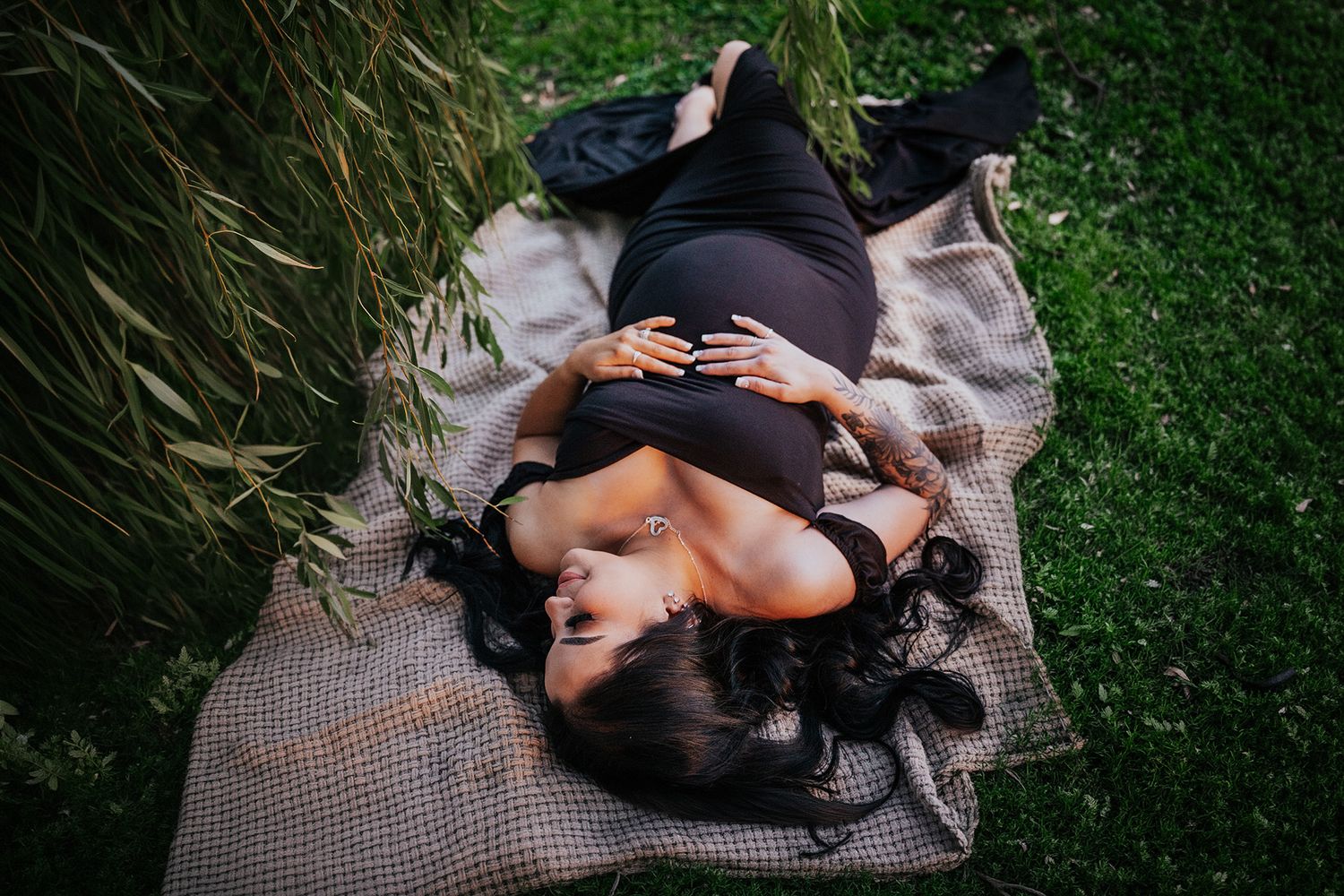 Pregnant woman in black dress lying on a blanket outdoors, cradling her baby bump surrounded by tall green grass.