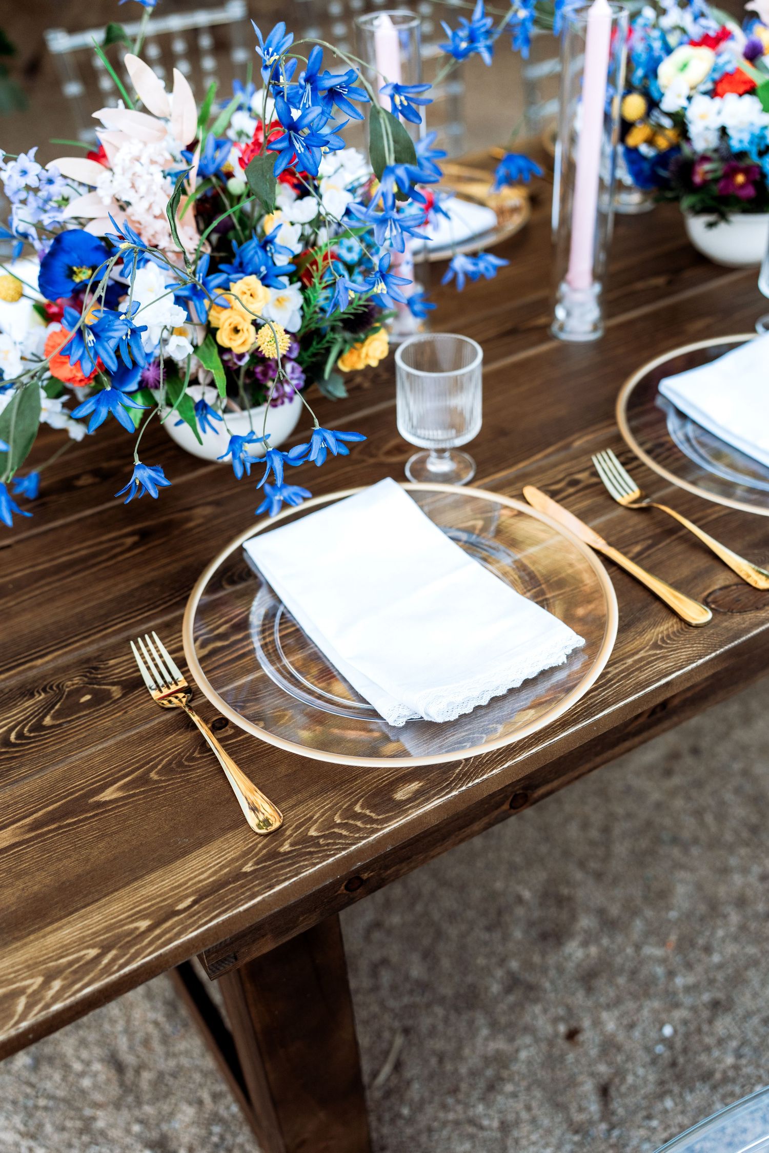 Clear charger plate with white napkin and gold flatware beside vibrant blue and multicolor floral centerpiece.