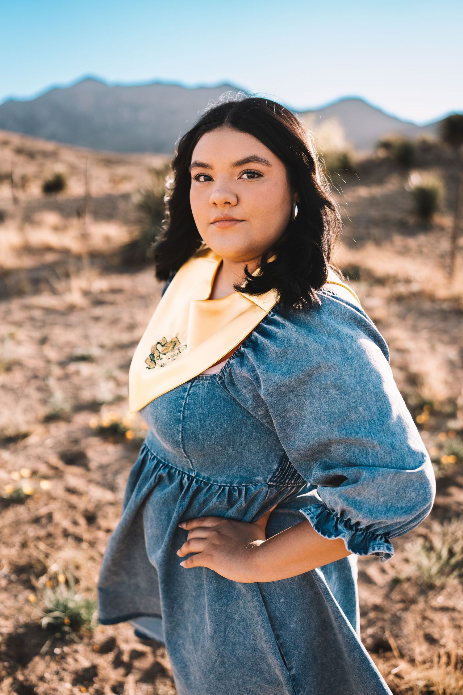 Young woman in denim dress and gold graduation stole posing in a sunlit desert landscape.