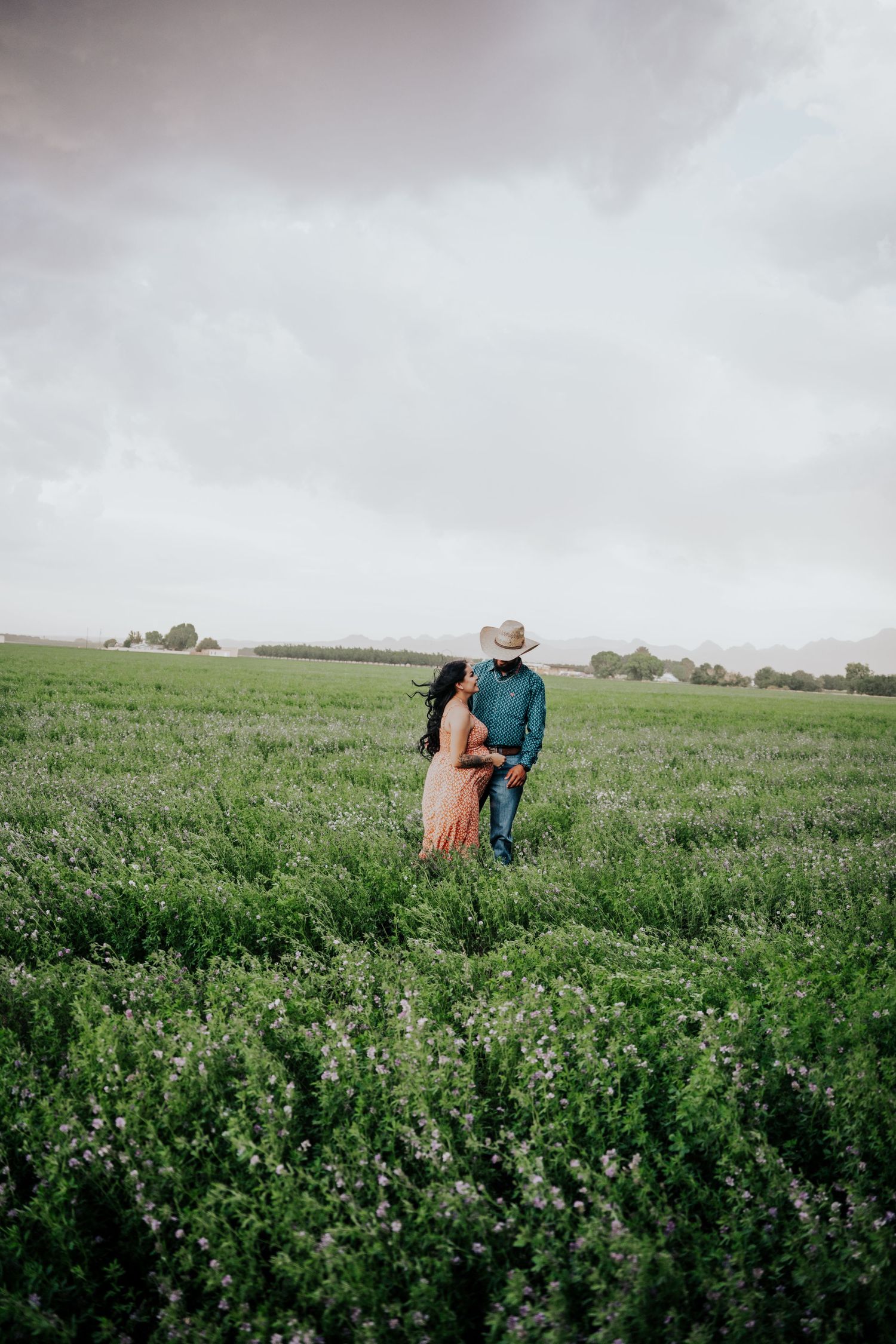 Pregnant couple standing together in a green field, woman in orange dress looking up at man in cowboy hat.