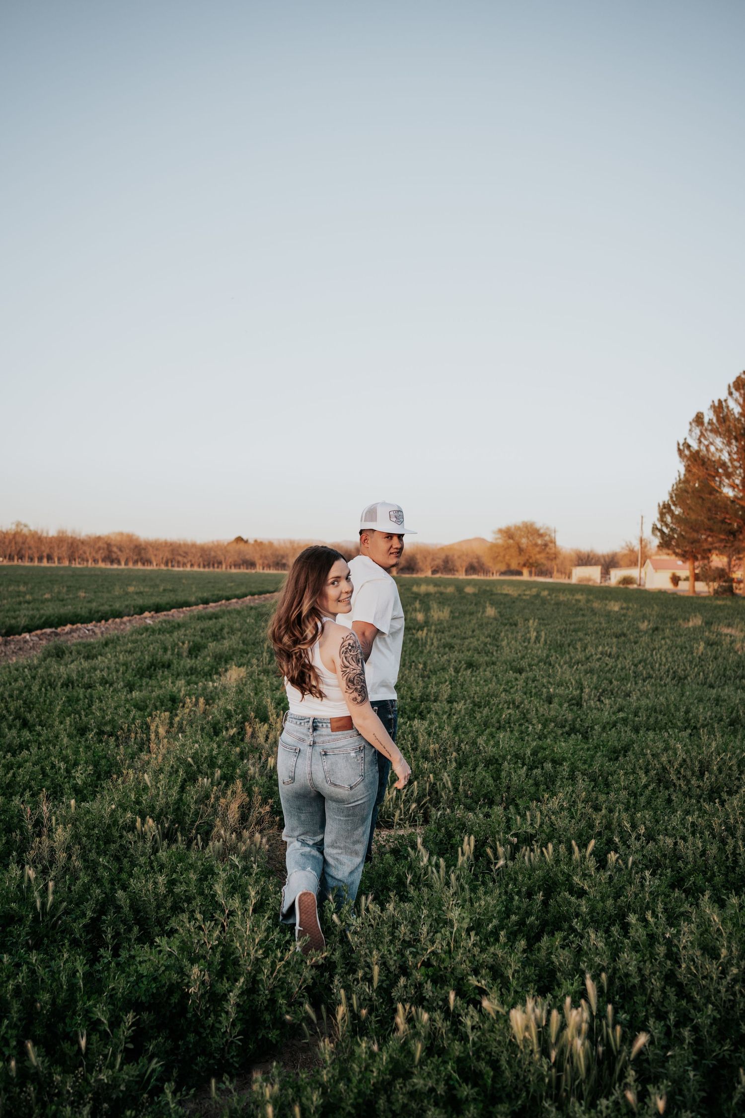 Couple standing in a green field at golden hour, woman with tattoo smiling back at camera.