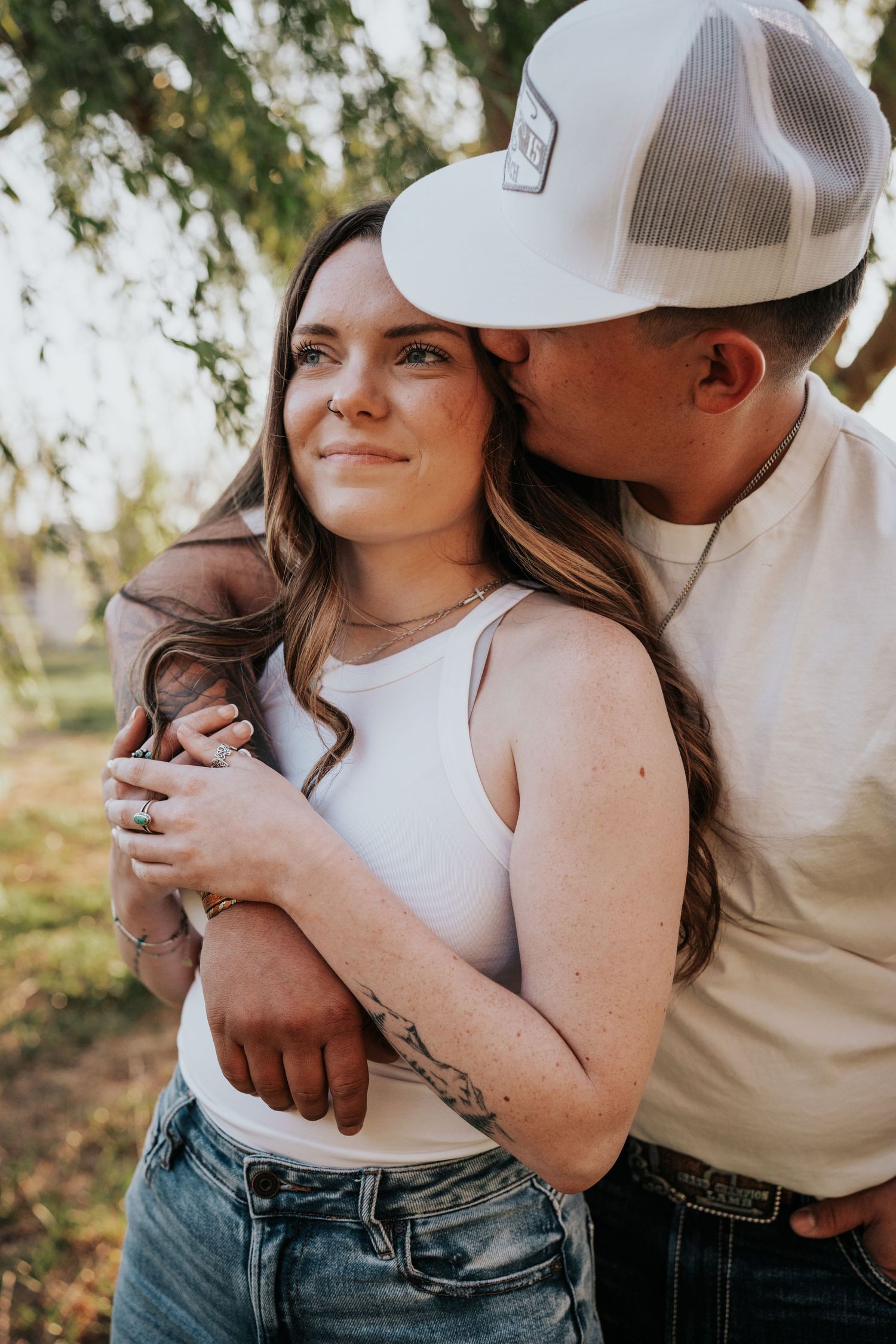 Couple embracing outdoors, man in white trucker hat kissing womans cheek, she smiles wearing white tank and jeans.