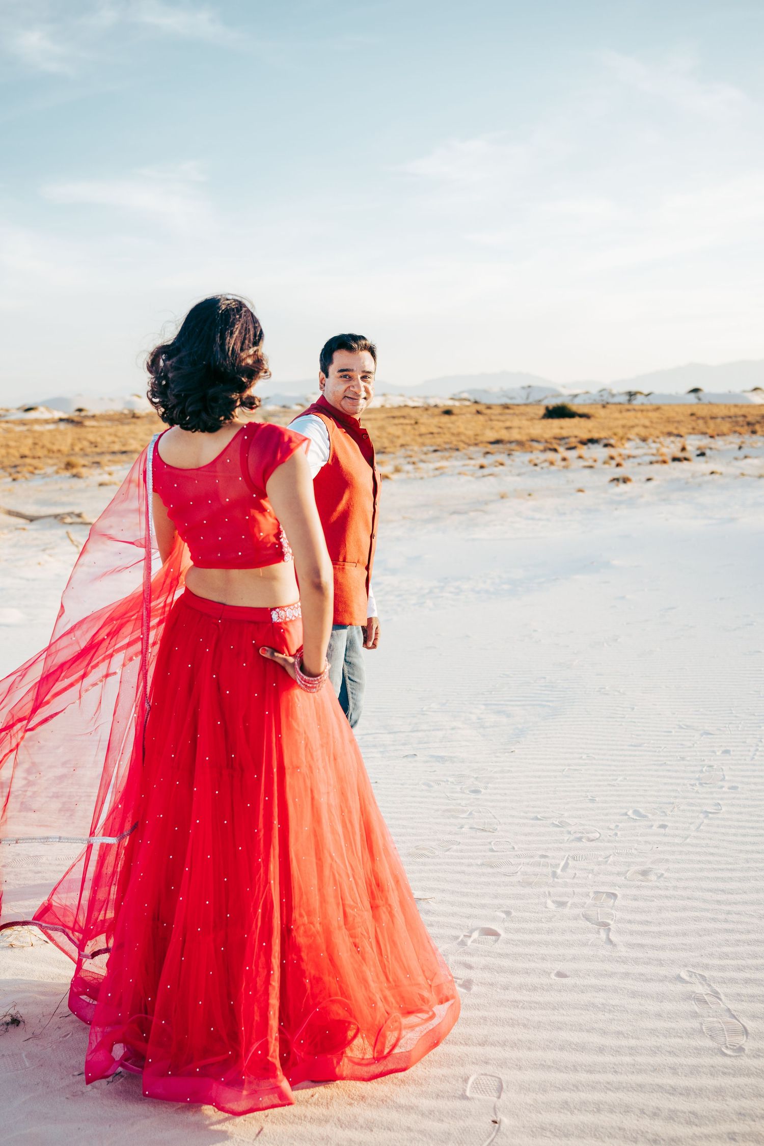 Woman in red lehenga with flowing dupatta stands with man in red vest on white desert sands.