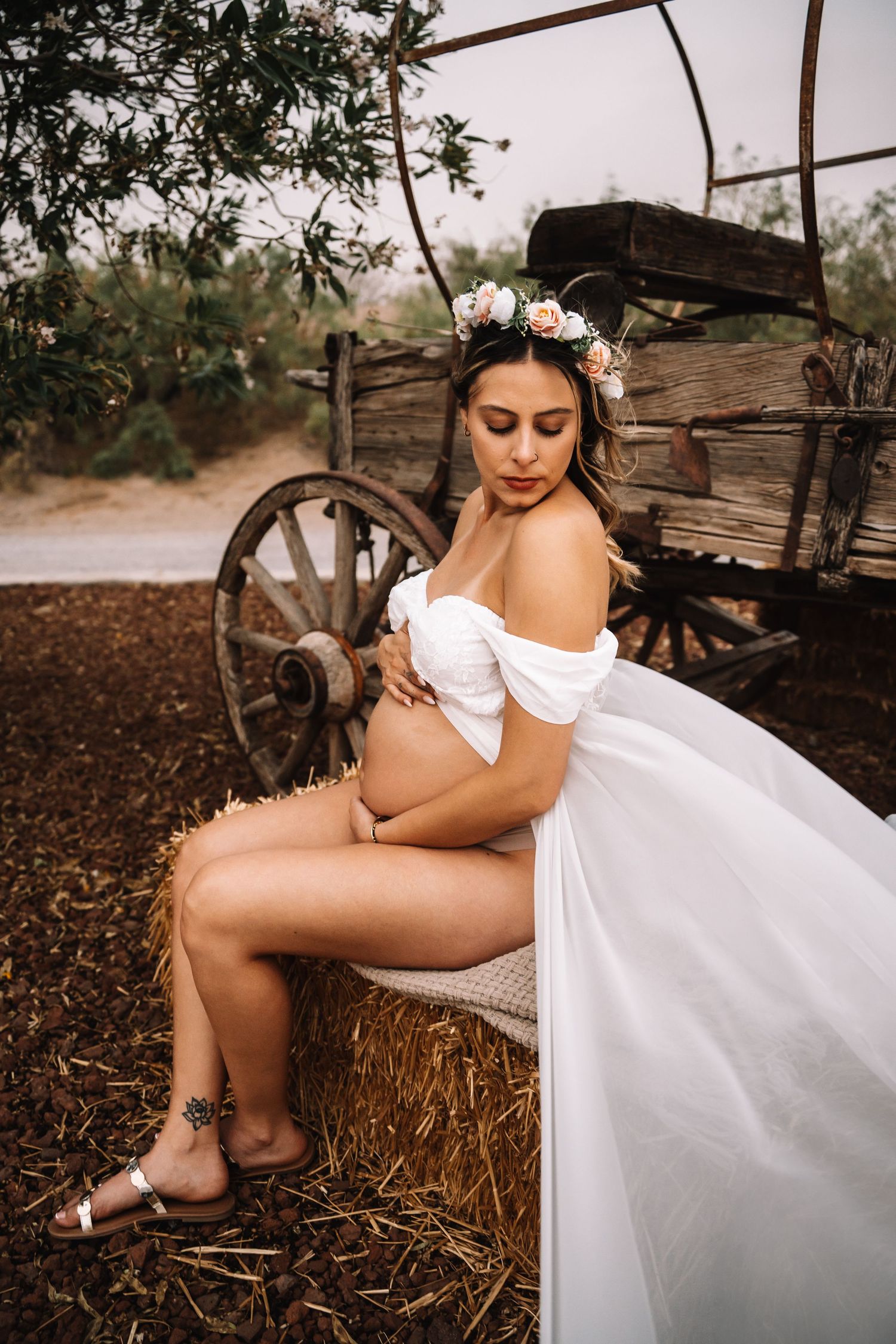 Pregnant woman in white off-shoulder gown and floral crown sits on hay bale beside rustic wooden wagon.