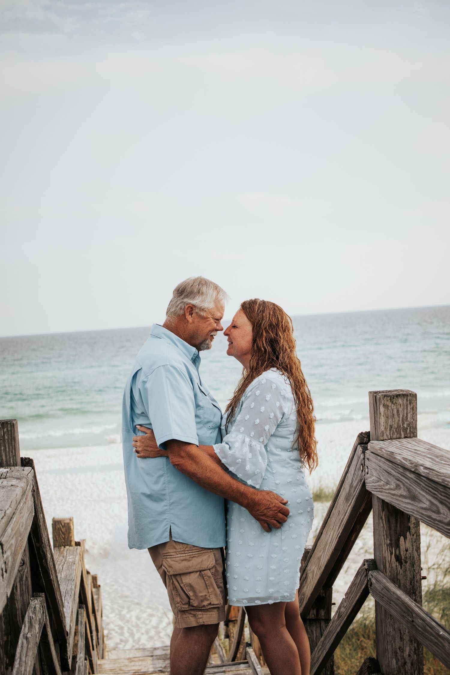 Older couple embracing and touching foreheads romantically on a beach boardwalk with ocean views.