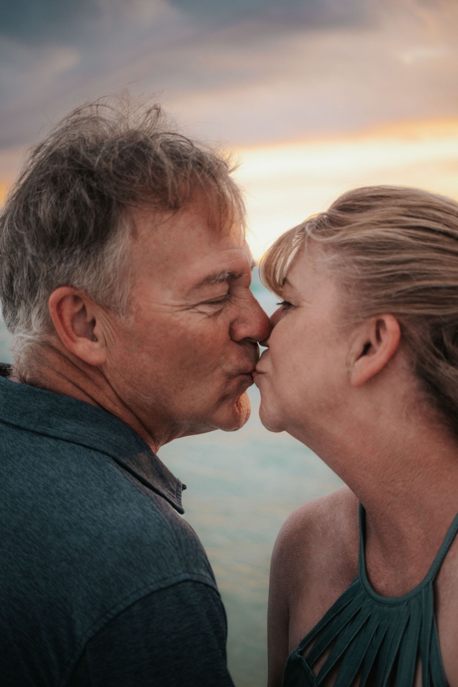 Close-up of mature couple sharing a tender kiss outdoors with a colorful sunset sky behind them.