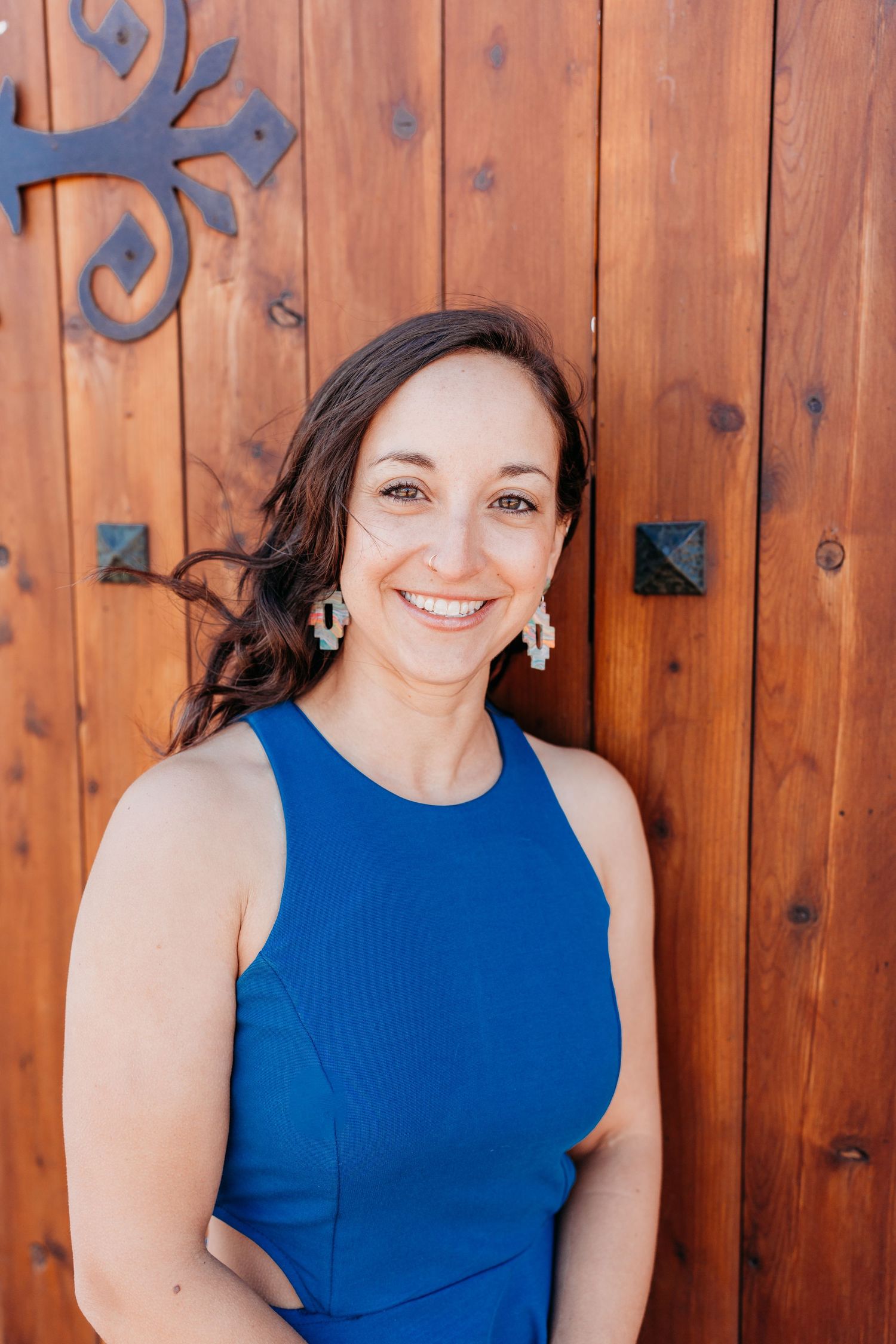 Smiling woman in blue dress leaning against wooden door with decorative ironwork.