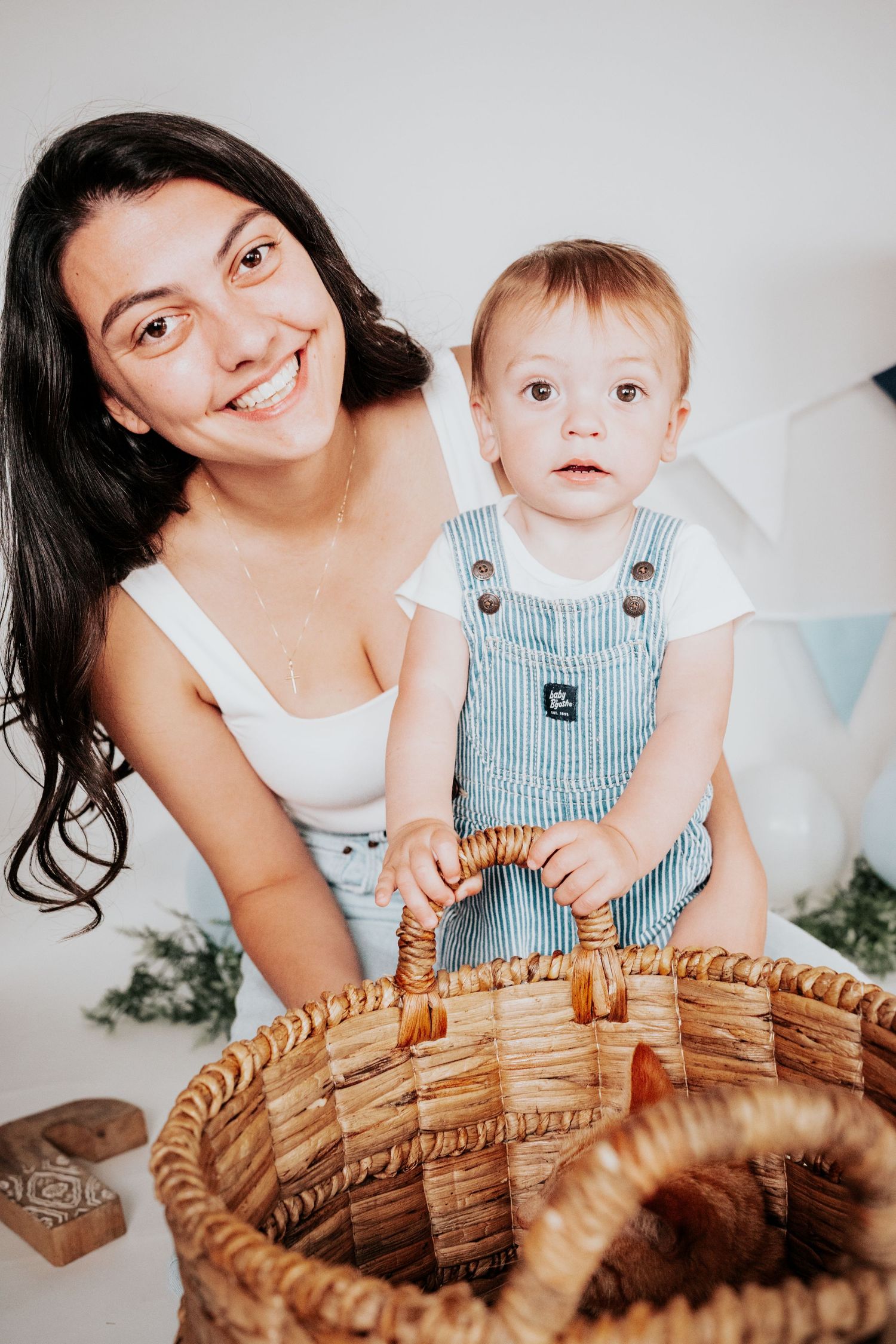 Smiling brunette mother posing with her toddler boy in striped overalls behind a large wicker basket.