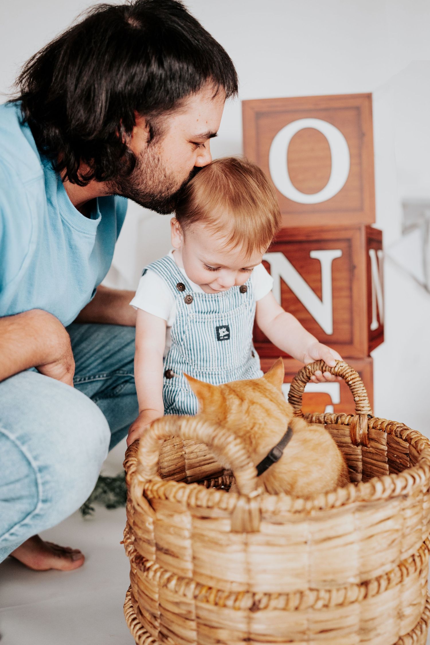 Bearded father kissing red-haired baby while both pet an orange cat in a wicker basket indoors.