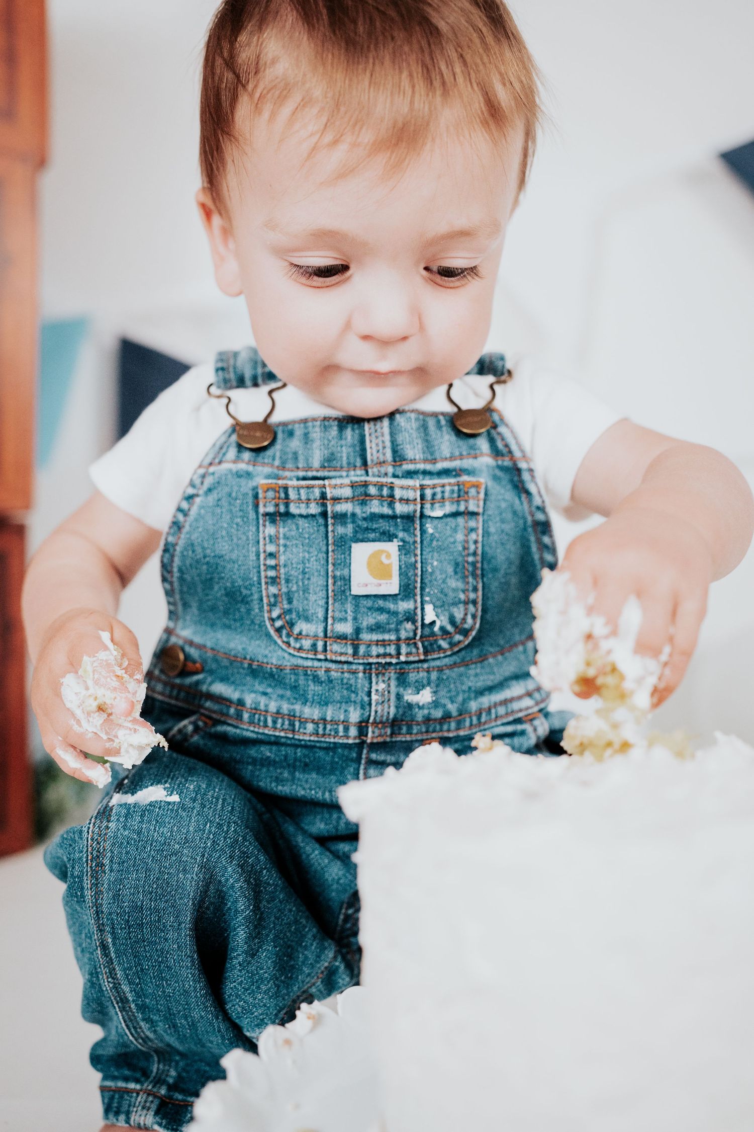 Baby boy in Carhartt denim overalls smiling while smashing white first birthday cake.
