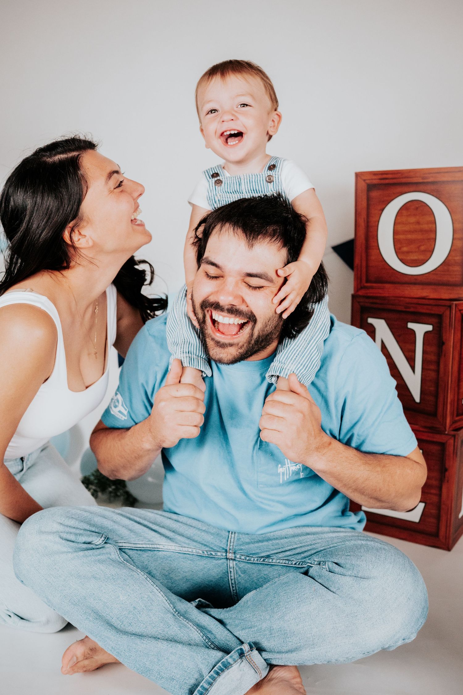 Joyful family with laughing baby on father's shoulders next to red ONE birthday blocks.