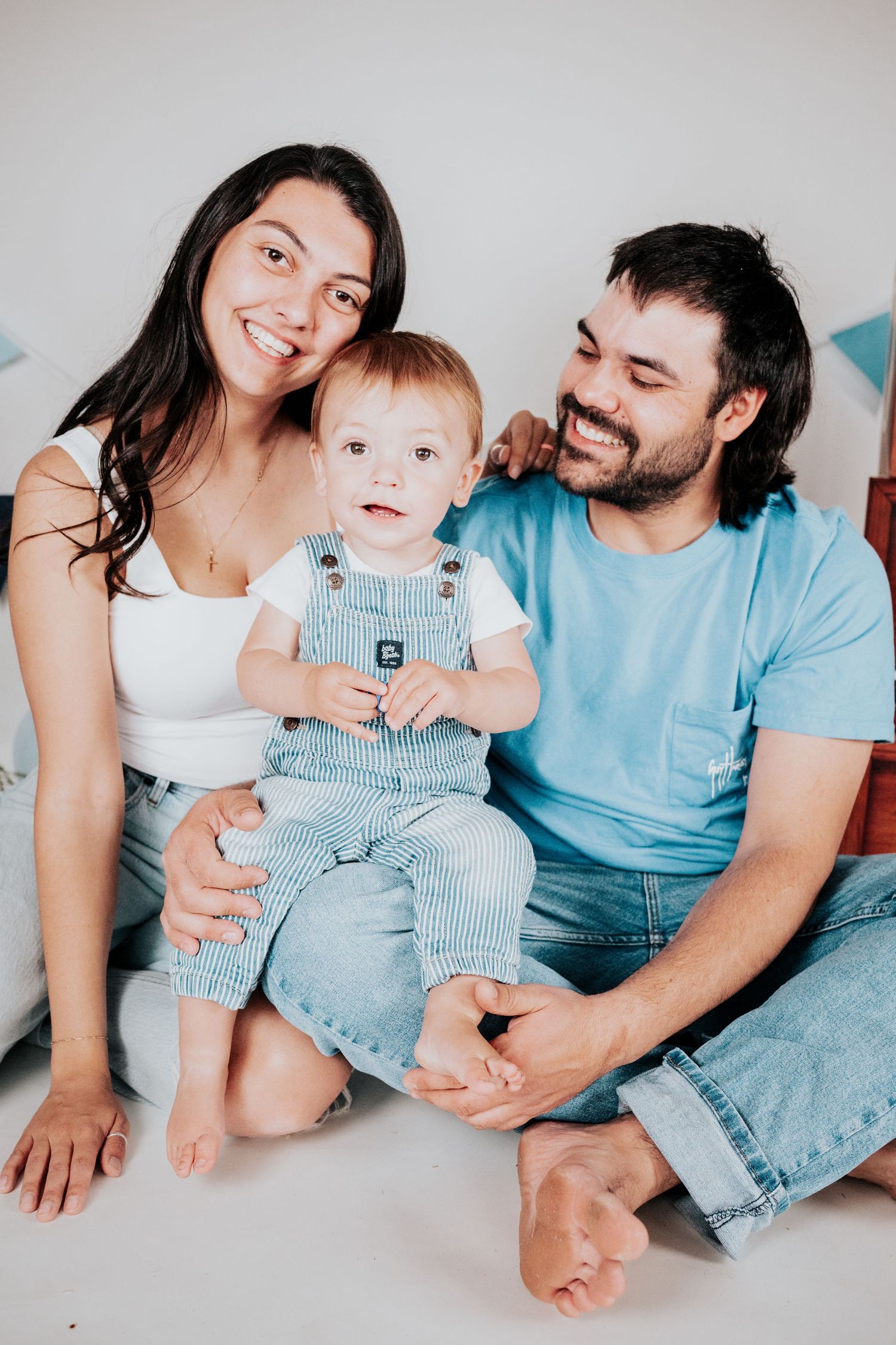 Loving family of three sitting together on floor, parents smiling at adorable baby in denim overalls.