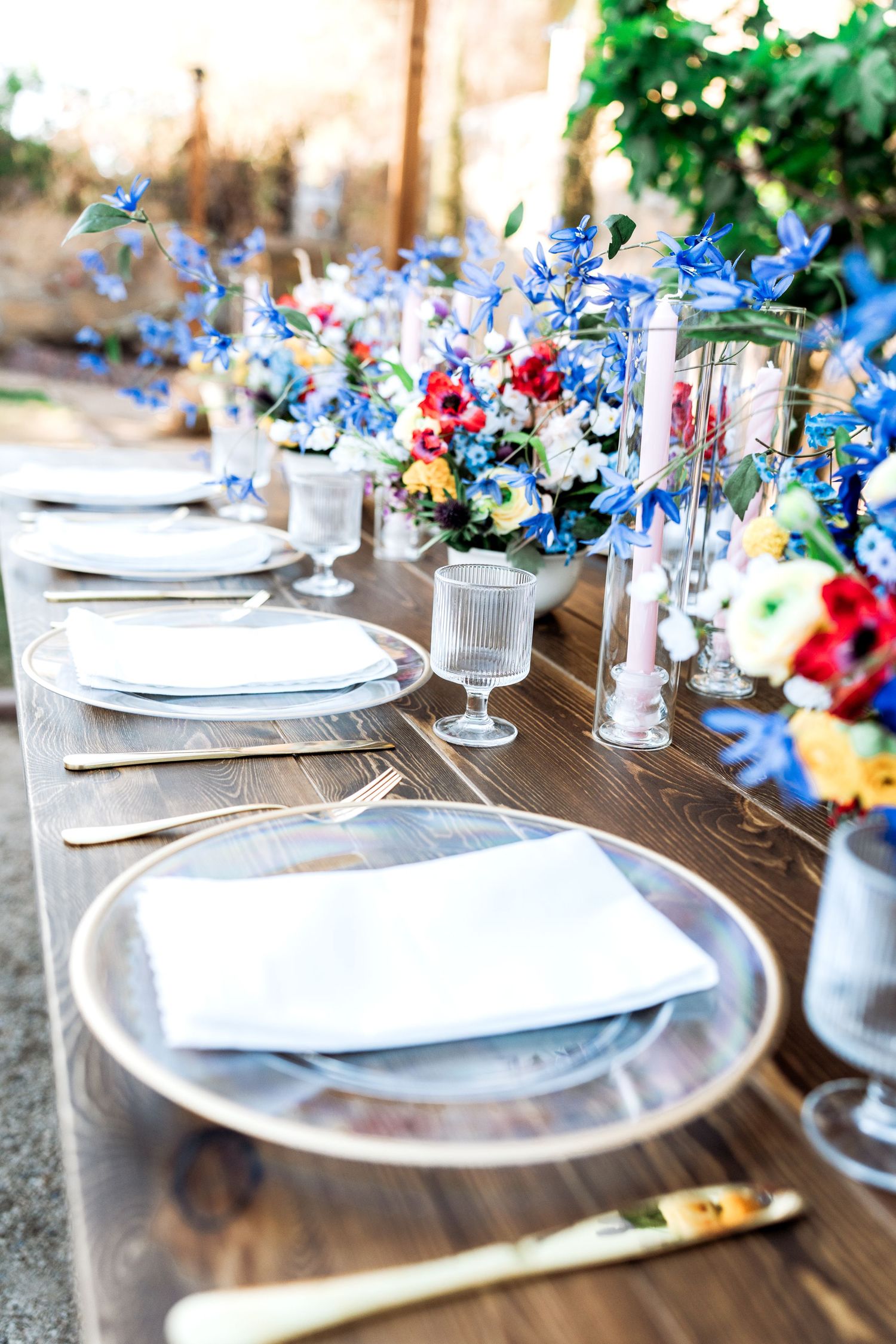 Long farm table with clear charger plates, gold flatware, and blue wildflower centerpieces for outdoor wedding.