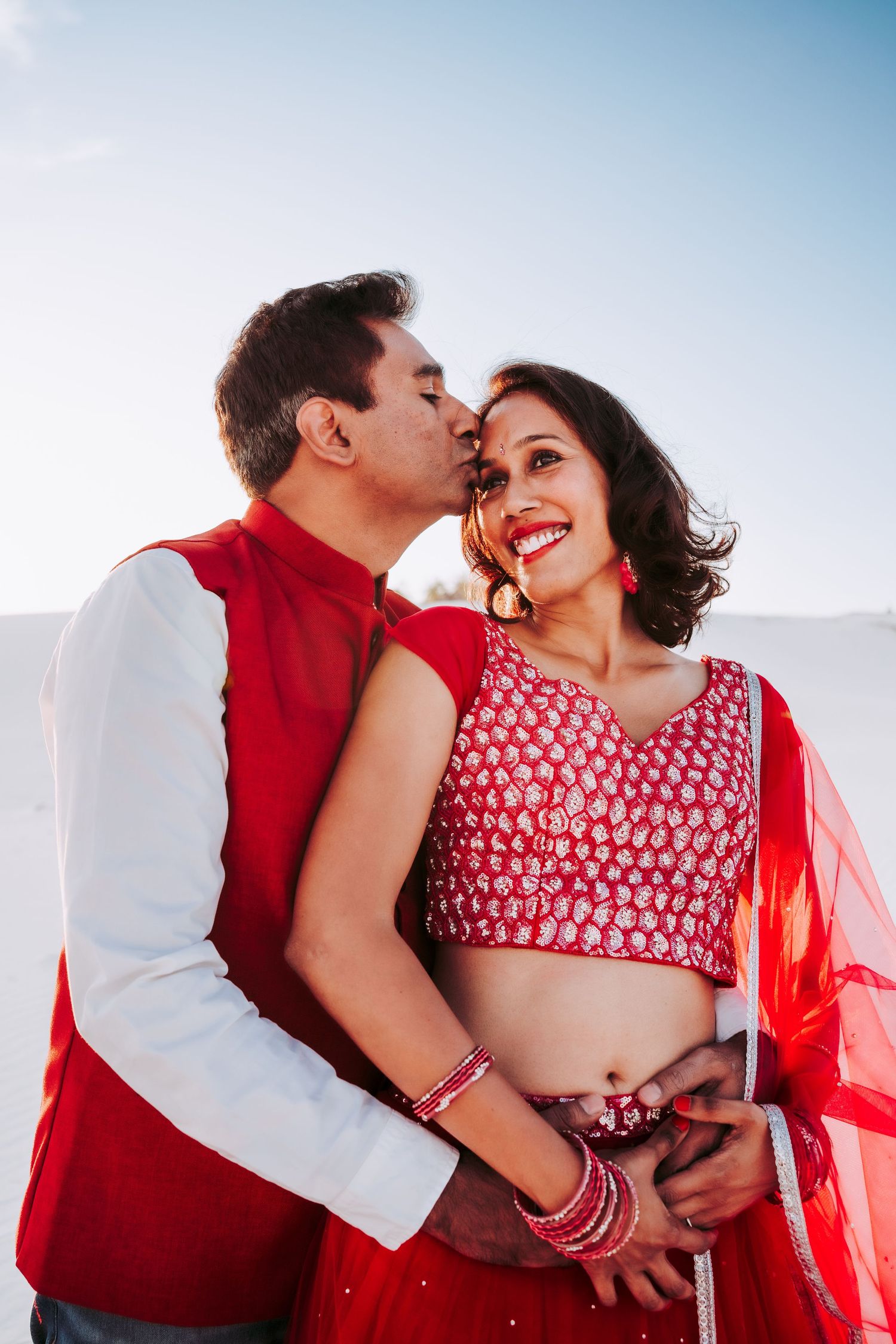 Couple in red traditional Indian attire sharing a romantic kiss on white sand dunes.