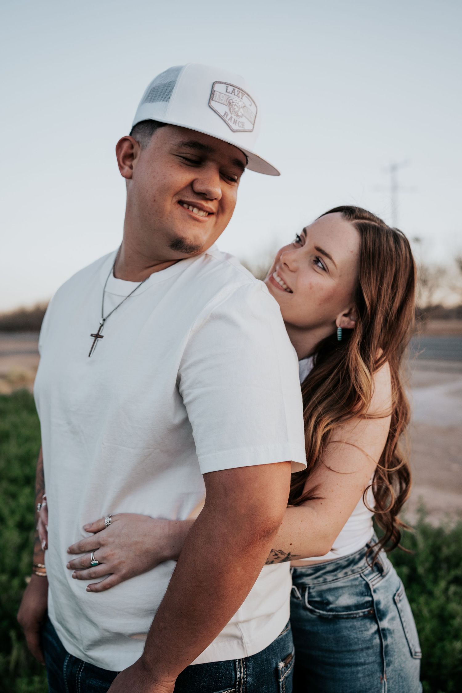 Woman with tattoo hugging man wearing white cap and cross necklace during outdoor couples photo session.