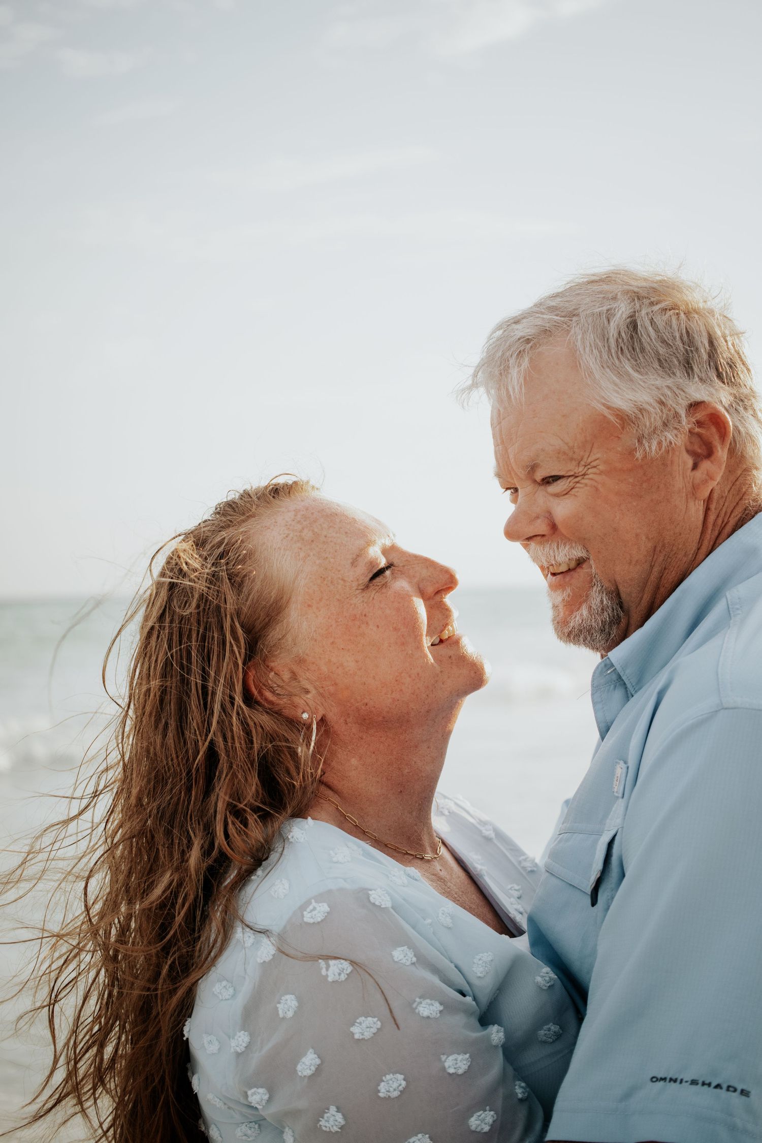 Joyful mature couple laughing and embracing closely on beach, man smiling broadly, woman with windswept hair.