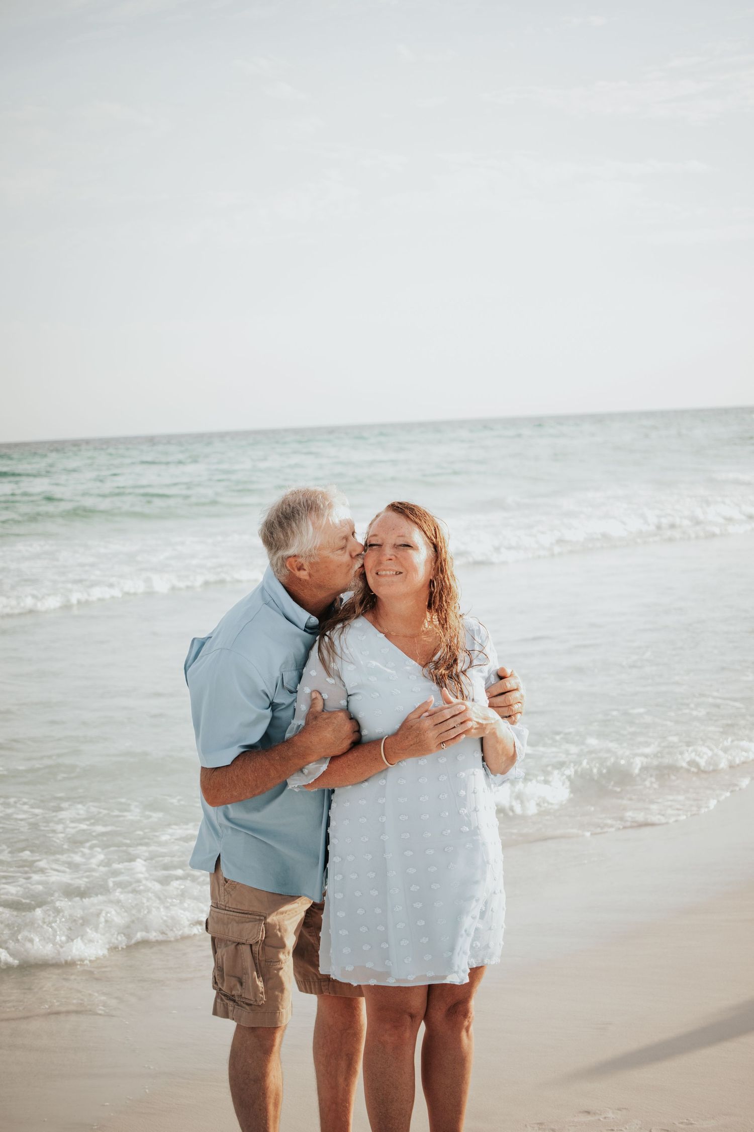 Older man kissing woman on the cheek while embracing on a sunny beach with gentle ocean waves.